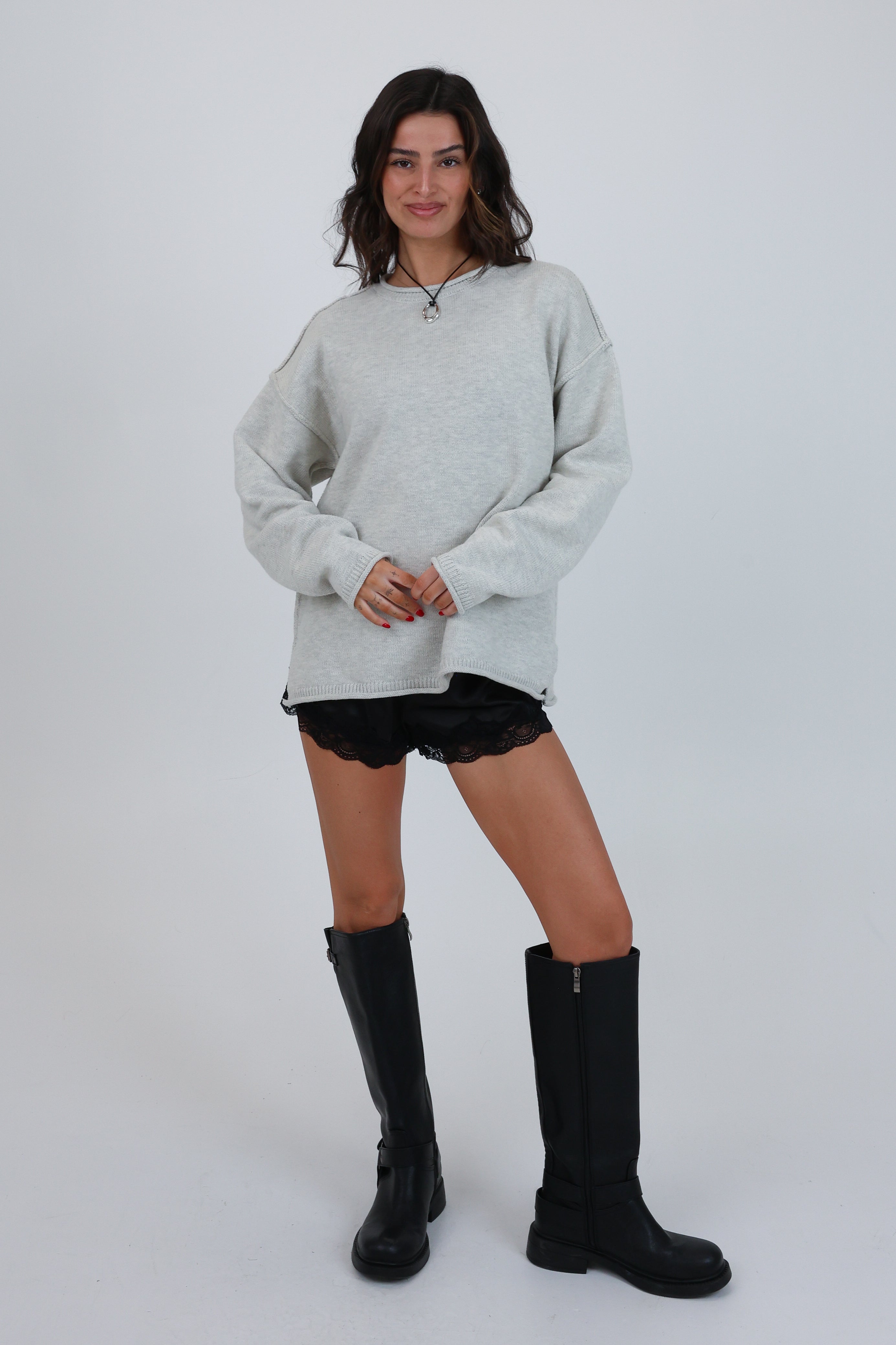 Woman wearing a light gray oversized sweater, black shorts, and black knee-high boots on a white background