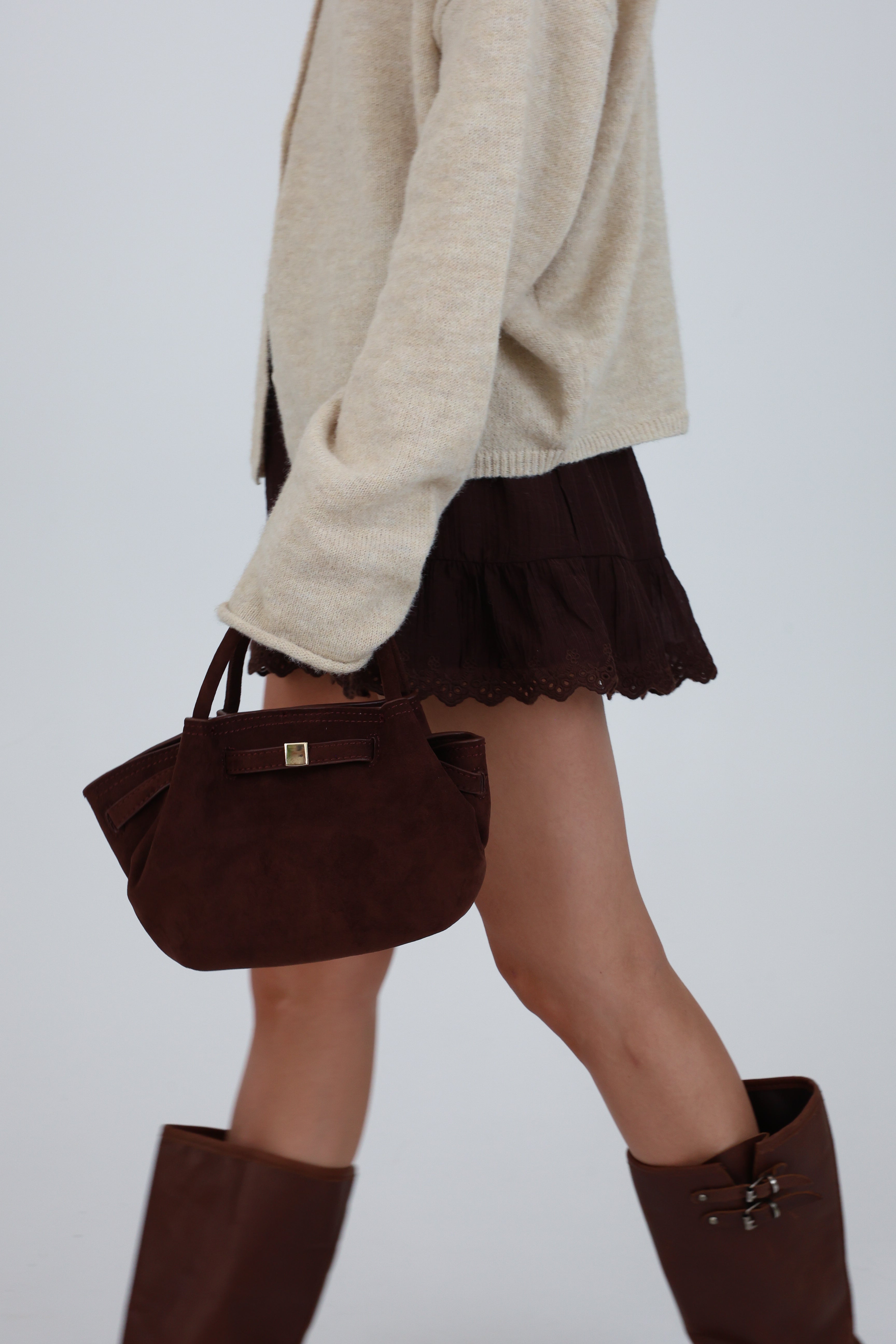 Person wearing a beige sweater, brown skirt, and brown knee-high boots holding a matching brown handbag on a plain background.