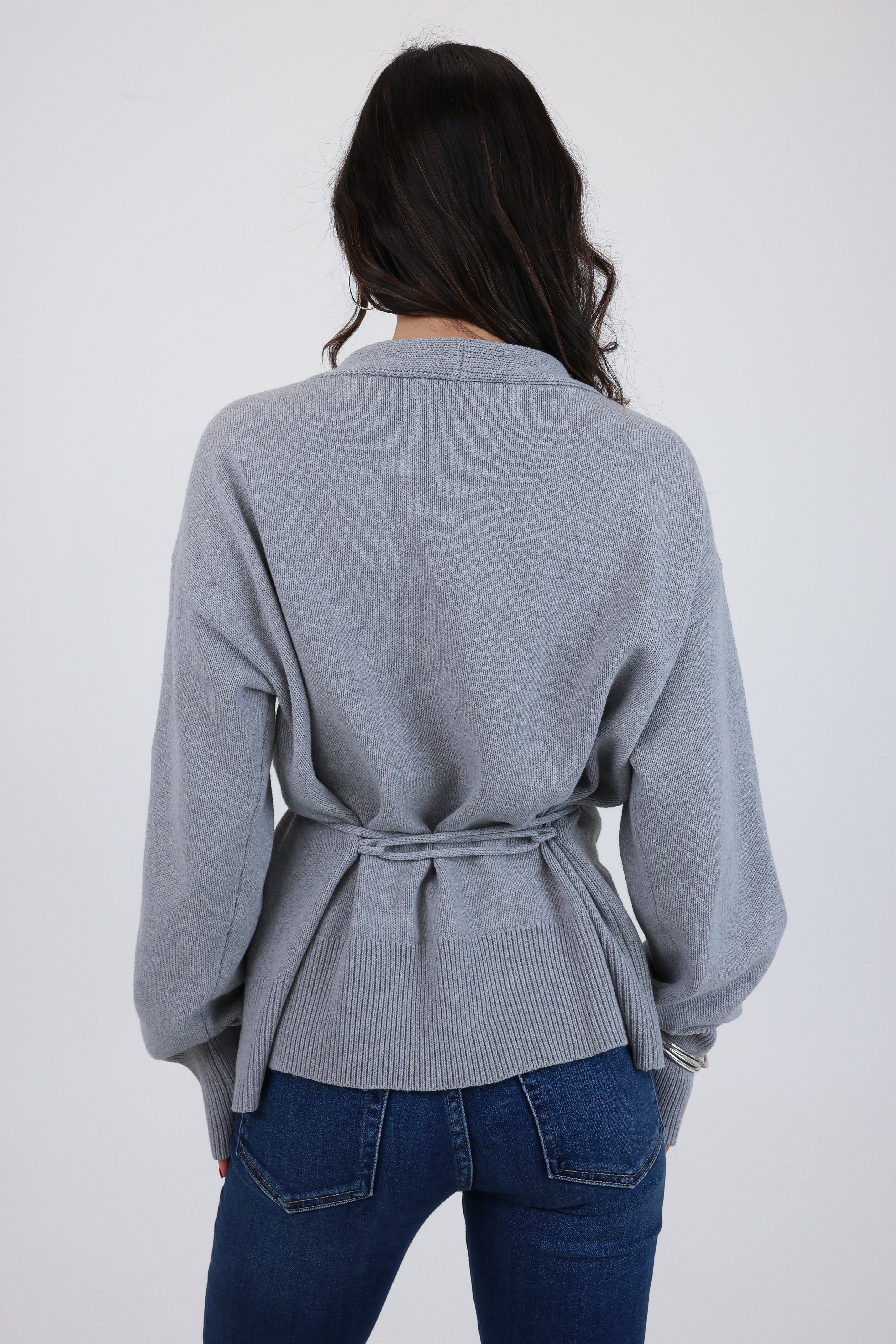 Person wearing a gray belted sweater over blue jeans on a white background