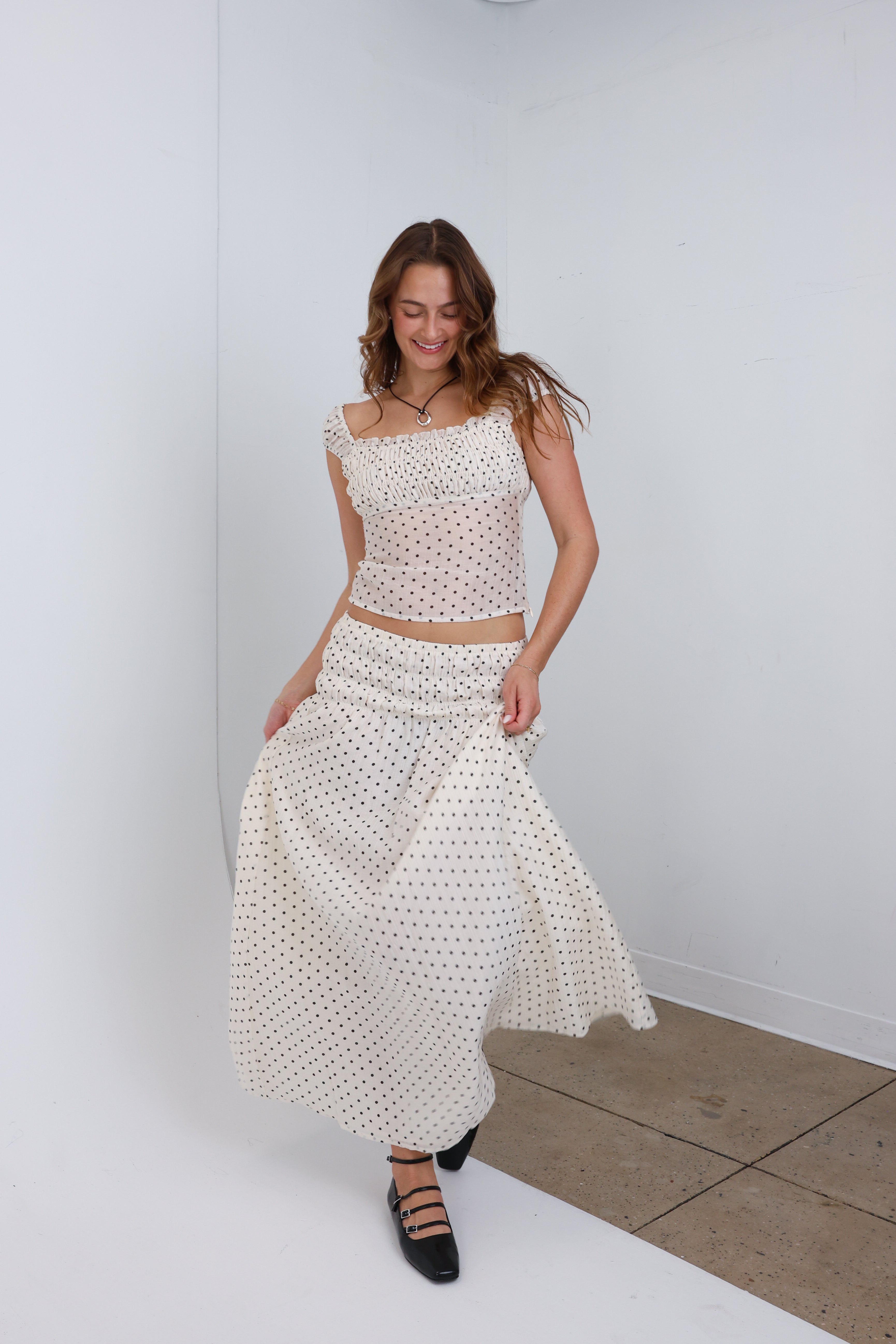 Woman wearing a white polka dot dress standing against a white wall.