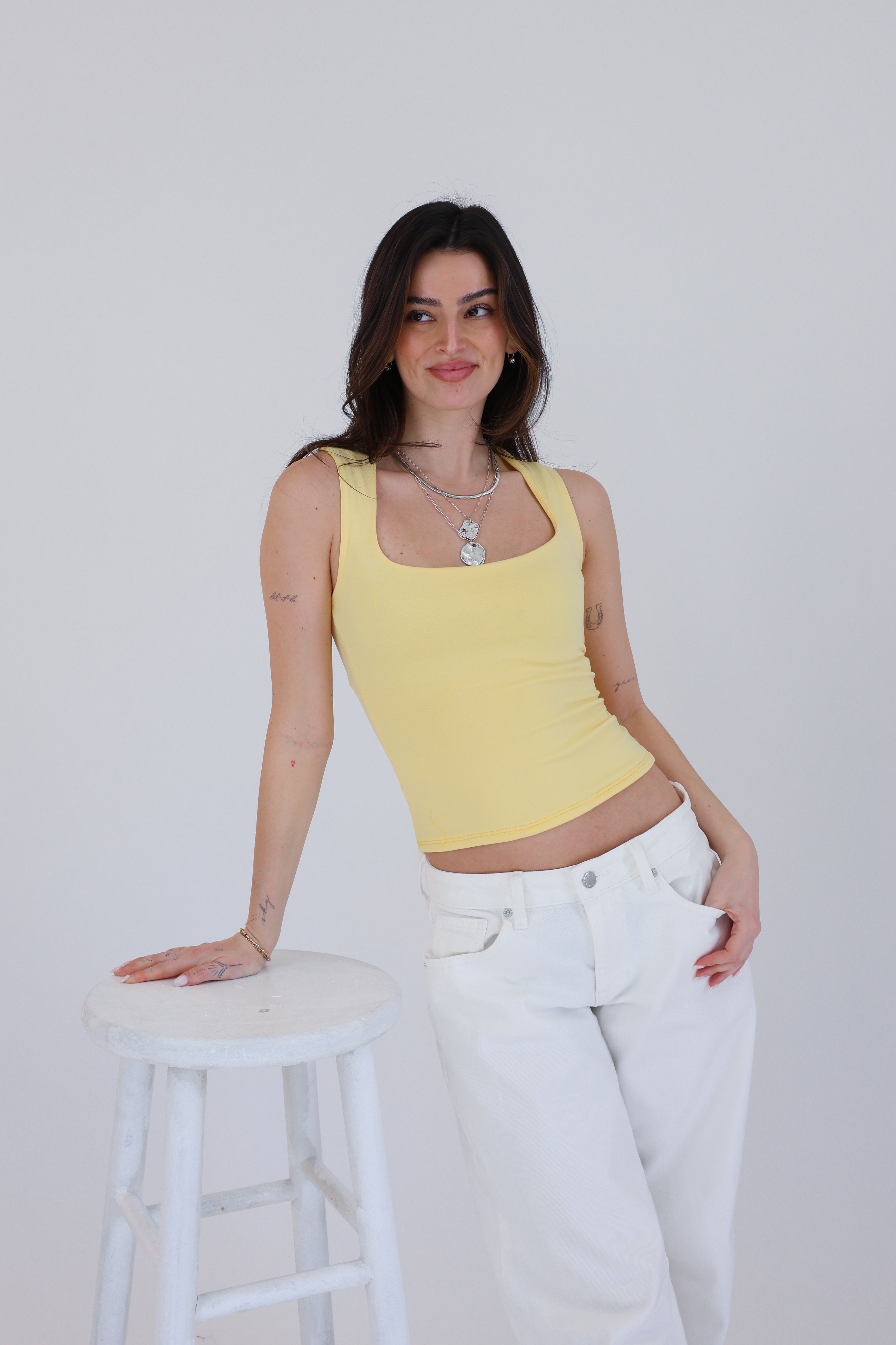 Woman wearing a yellow tank top and white pants standing on a white stool against a plain background