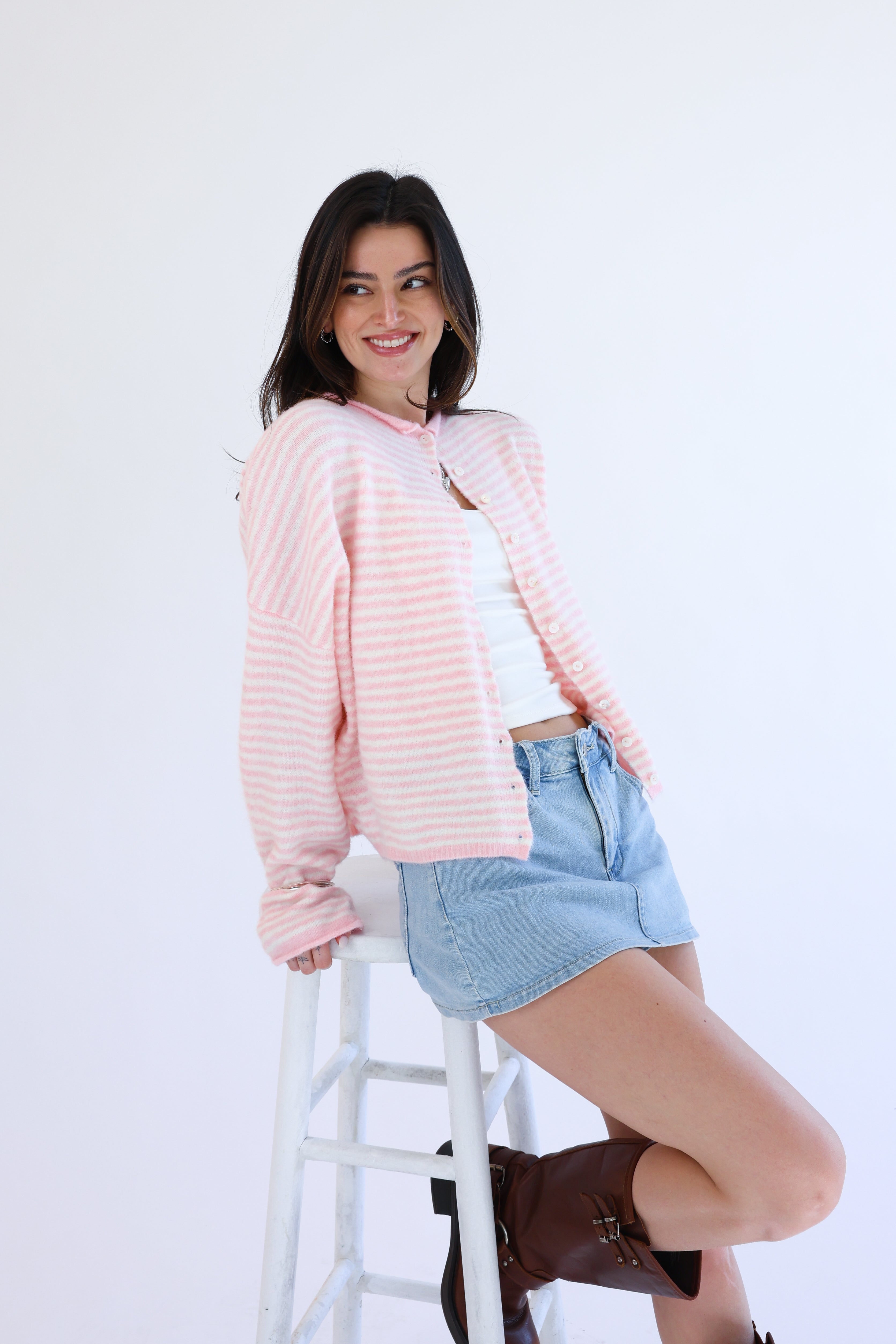 Woman wearing a pink textured cardigan over a white top and denim shorts, standing on a white stool against a white background.