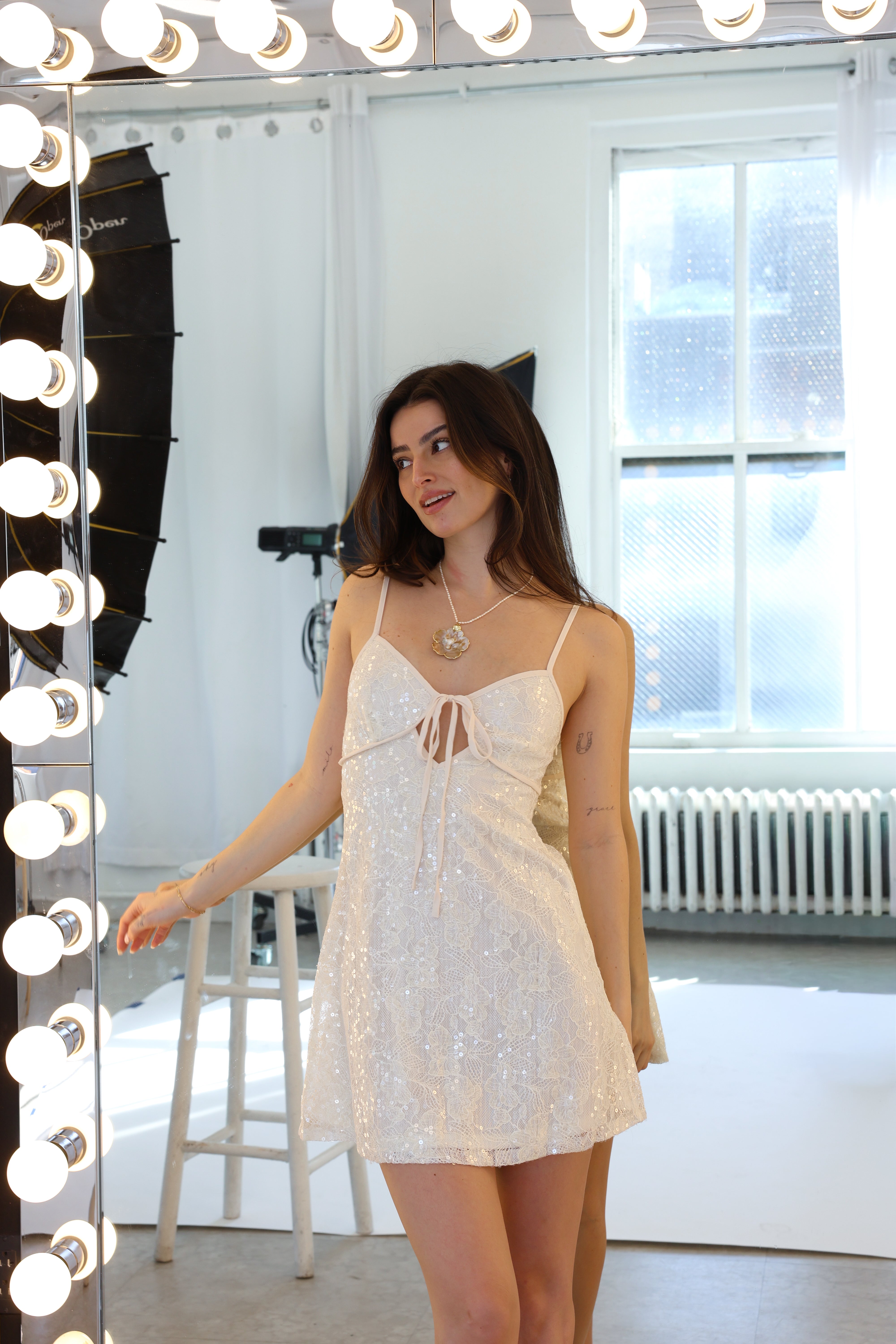 Woman wearing a white lace dress standing in front of a mirror with light bulbs.