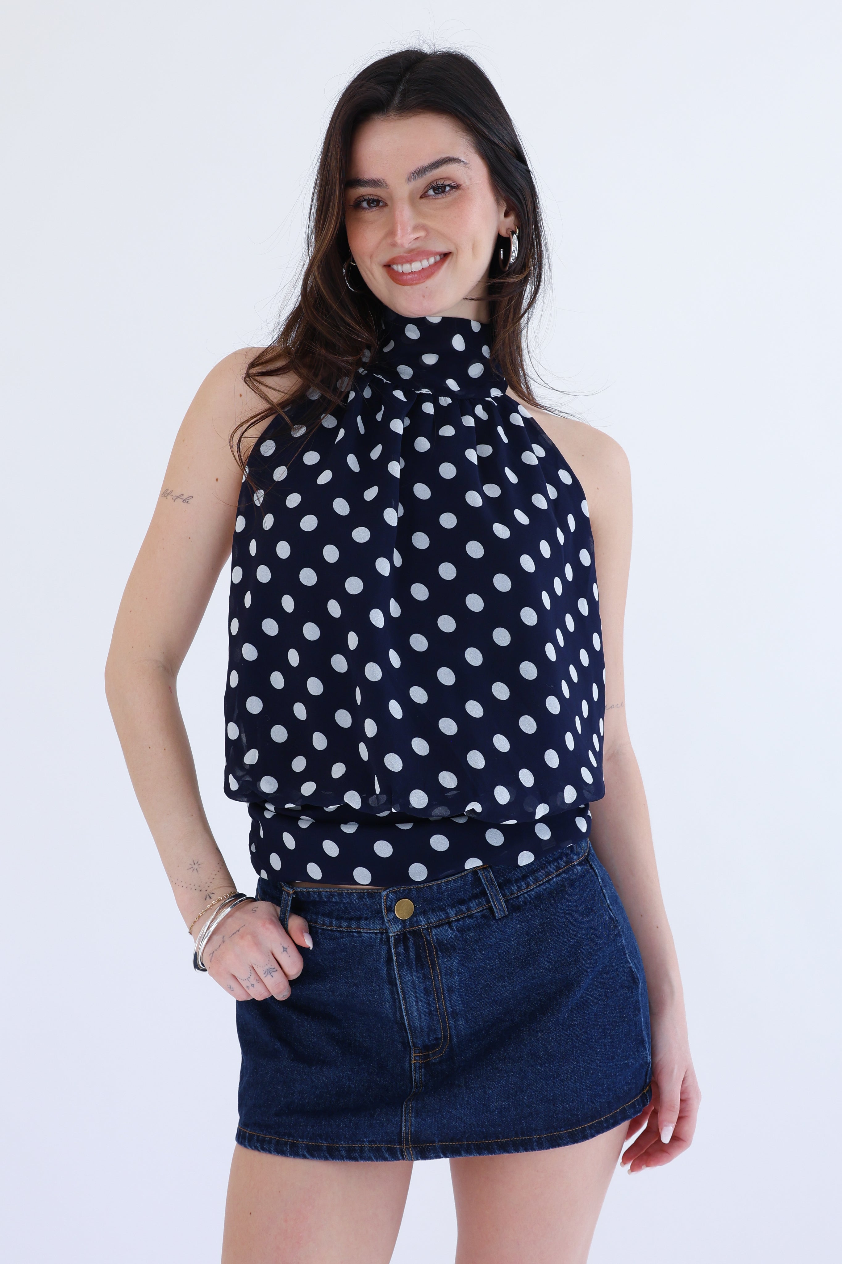 Woman wearing a navy blue sleeveless top with white polka dots and a denim skirt on a white background