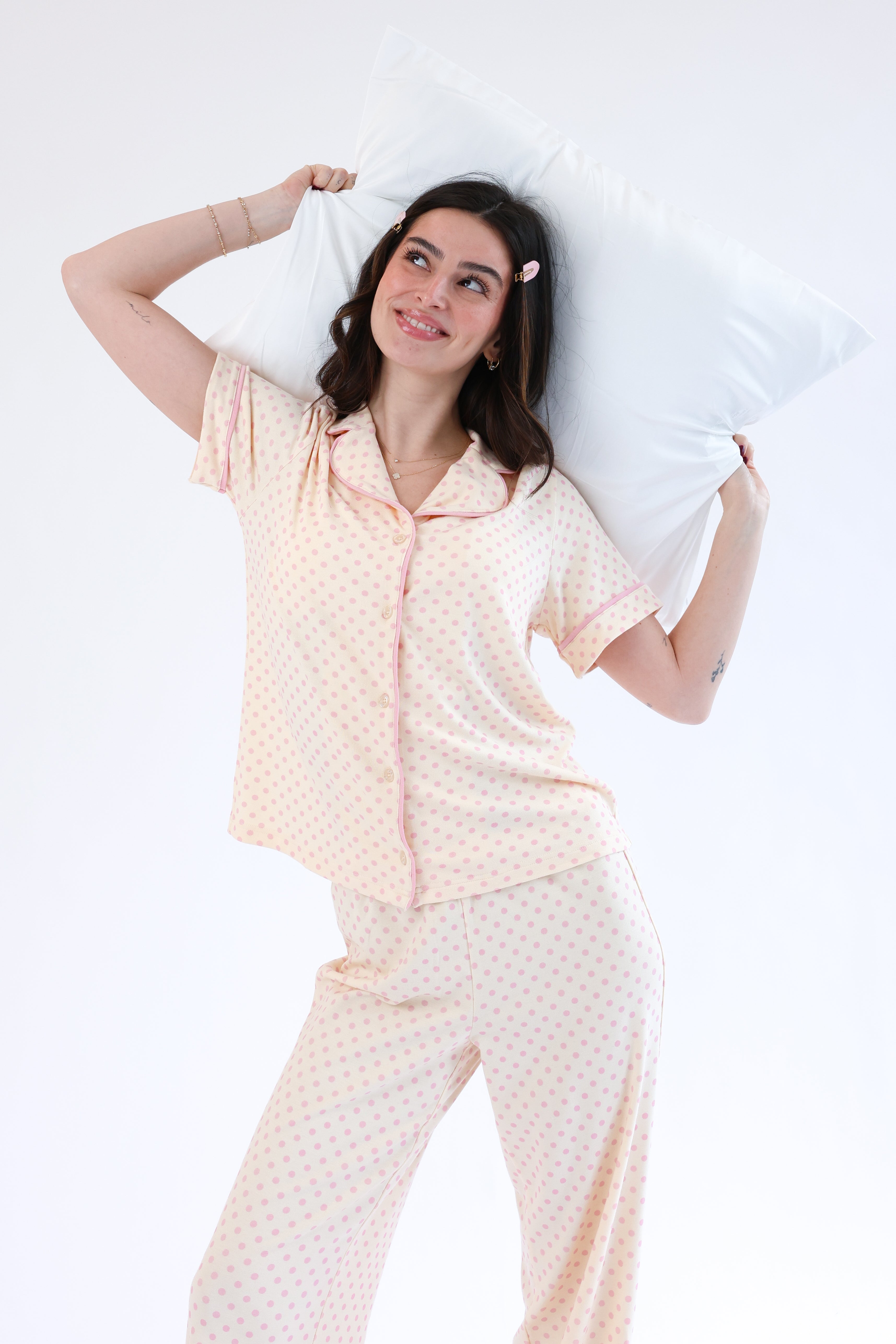 Woman wearing a light pink pajama set with a white pillow on a plain background