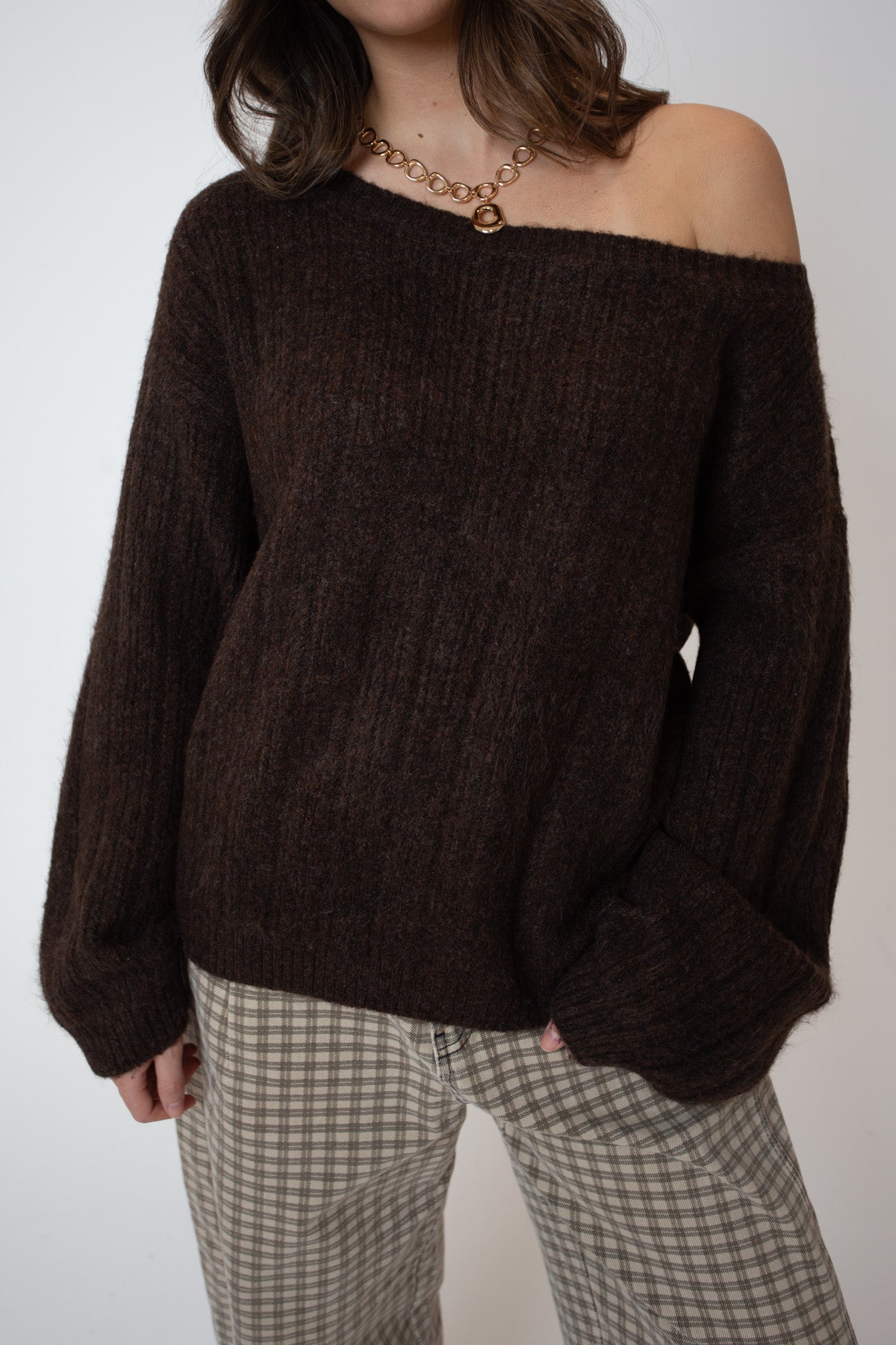 Person wearing a brown off-shoulder sweater and plaid pants on a white background