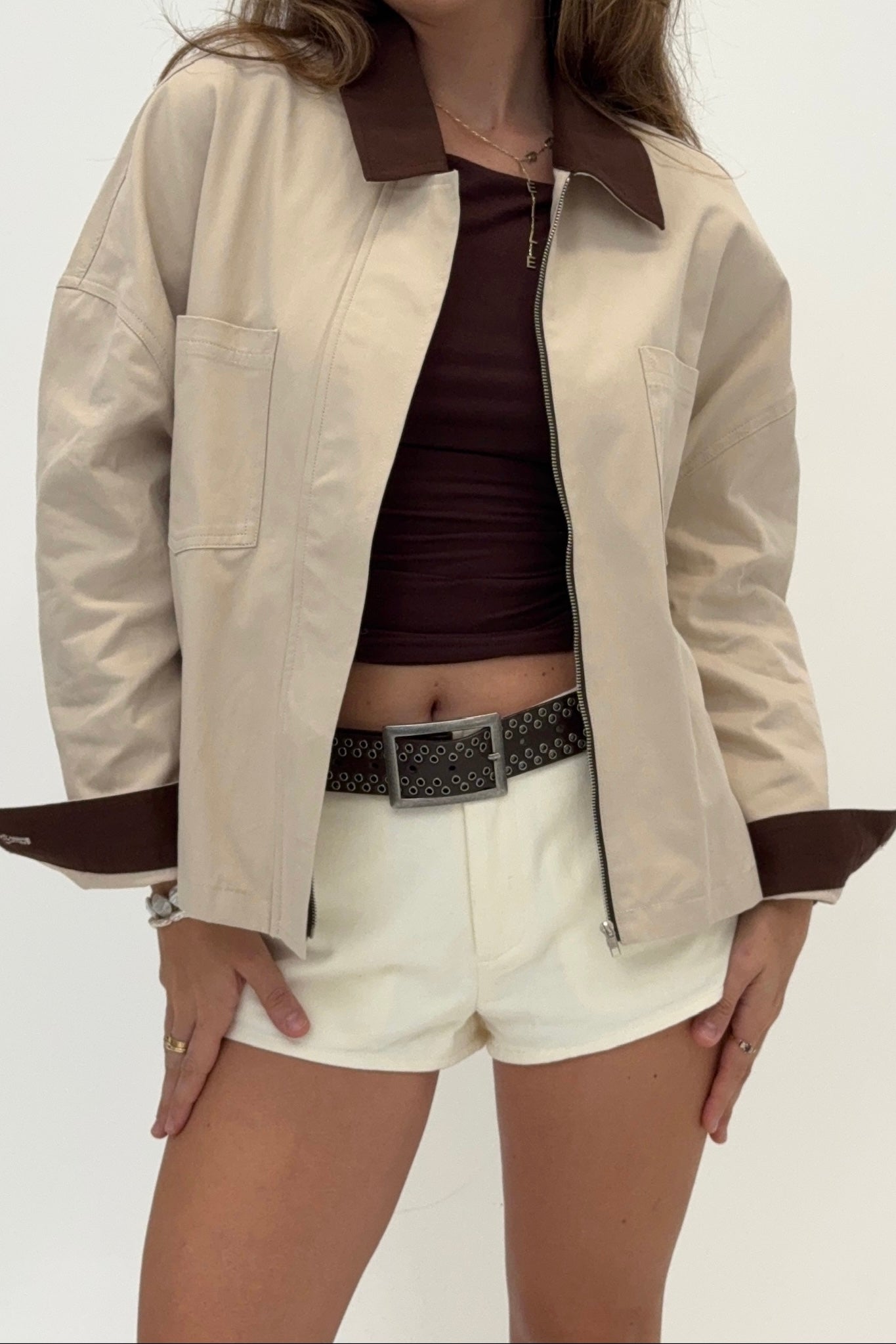 Person wearing a beige jacket with brown trim, brown top, and white shorts on a plain background
