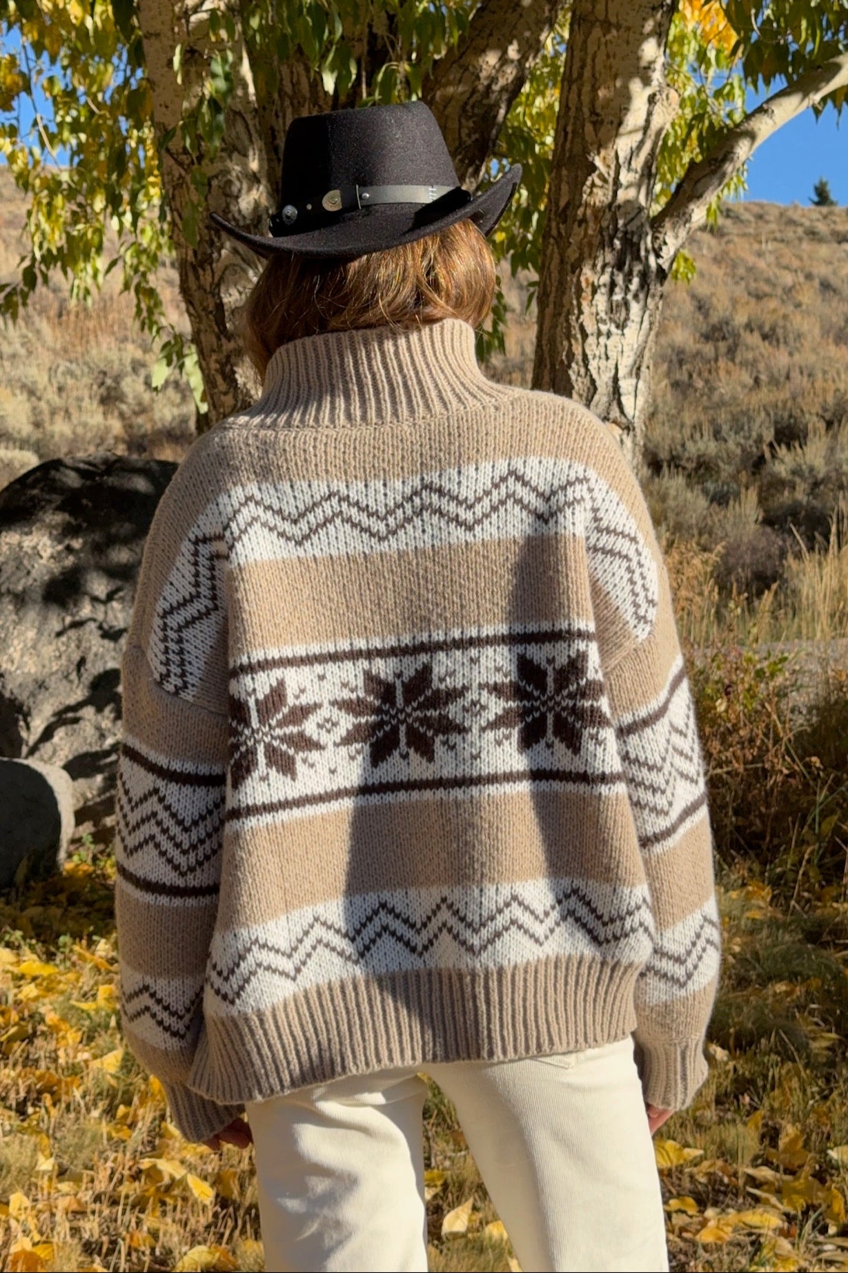 Person wearing a patterned sweater and cowboy hat in a natural setting with trees and grass.