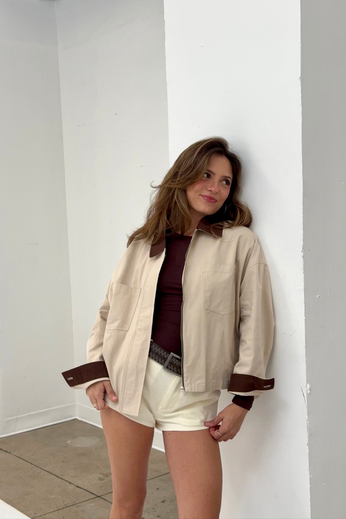 Woman wearing a beige jacket with brown accents against a white wall.