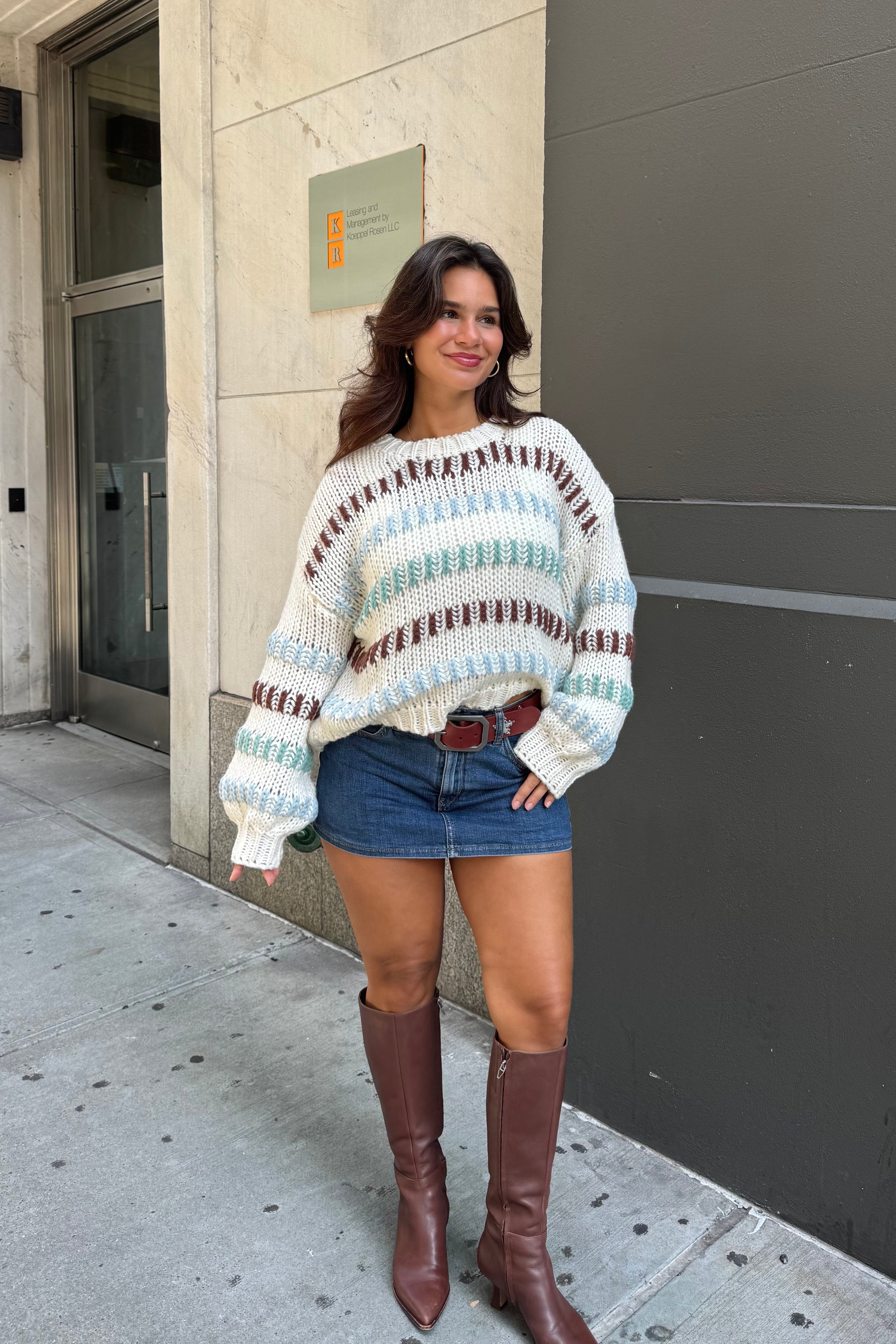 Woman wearing a colorful sweater and denim shorts standing on a sidewalk.
