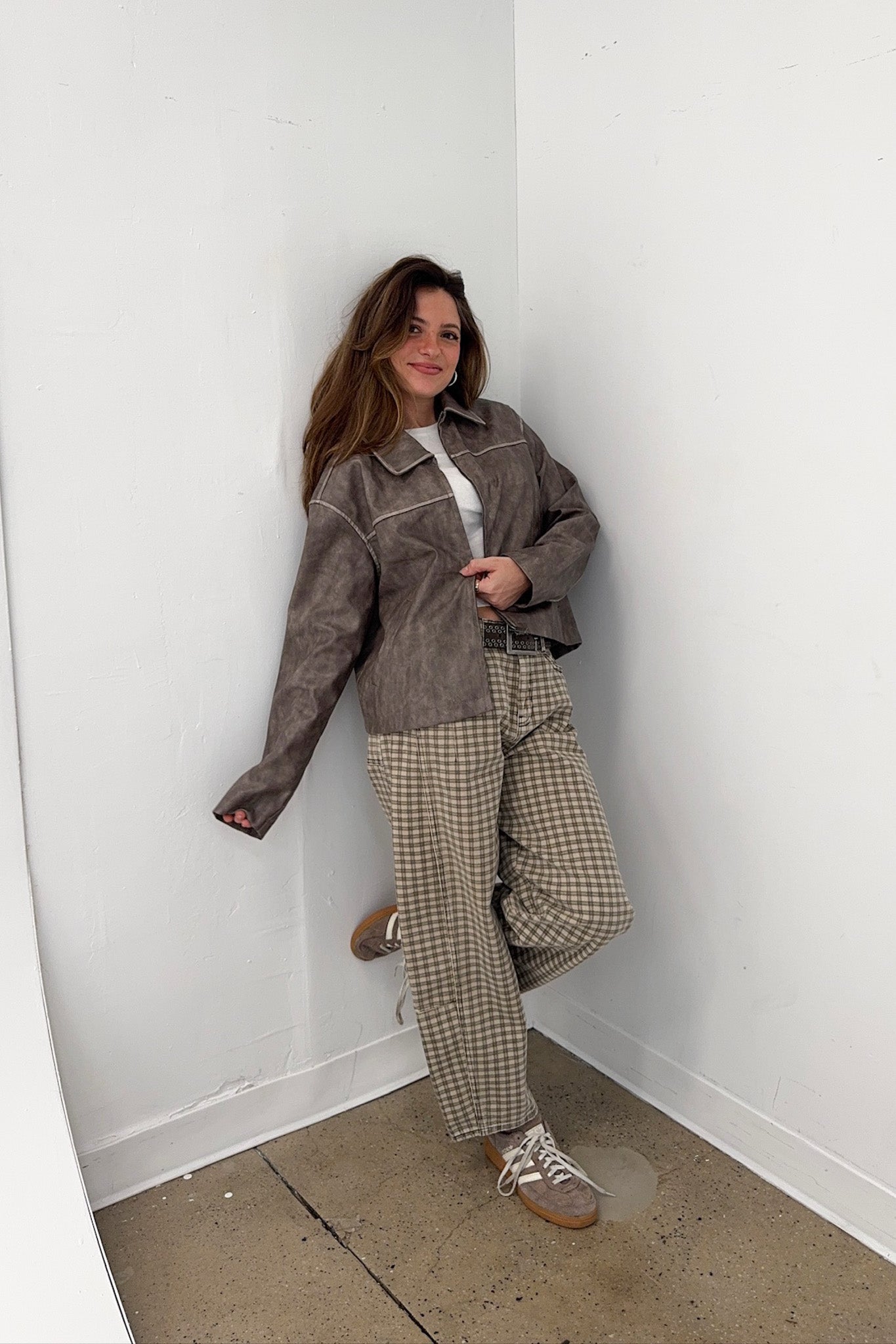 Woman wearing a brown jacket and plaid pants standing against a white wall.