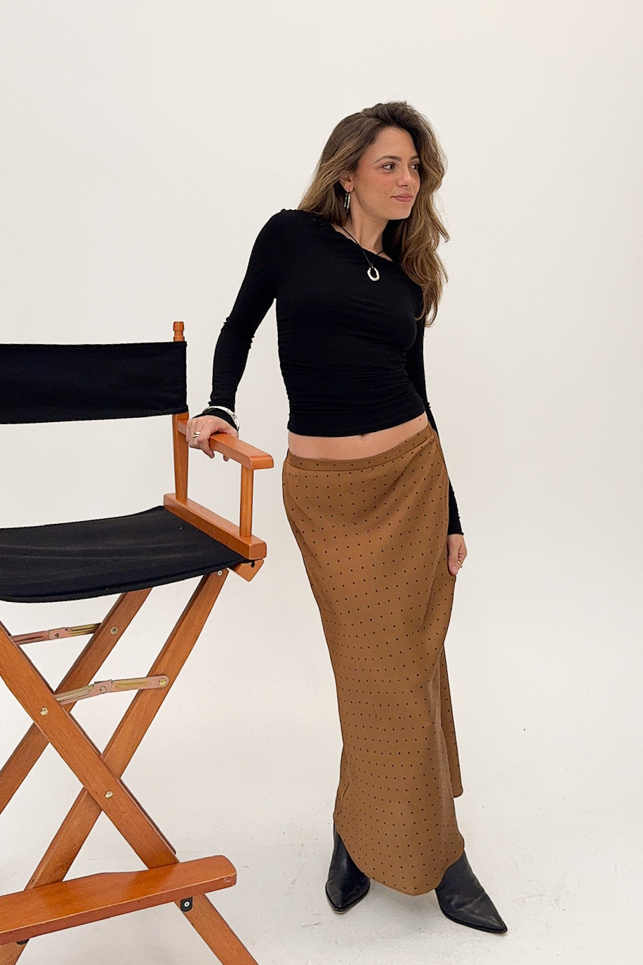 Woman in a black top and brown skirt standing next to a director's chair on a white background
