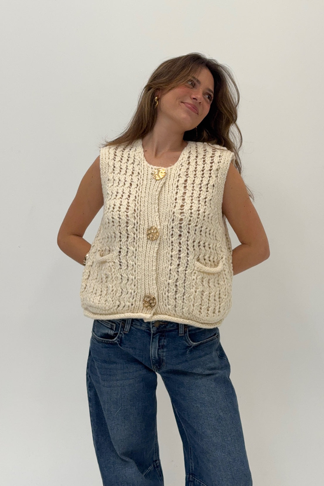 Woman wearing a beige knitted vest and blue jeans against a white background