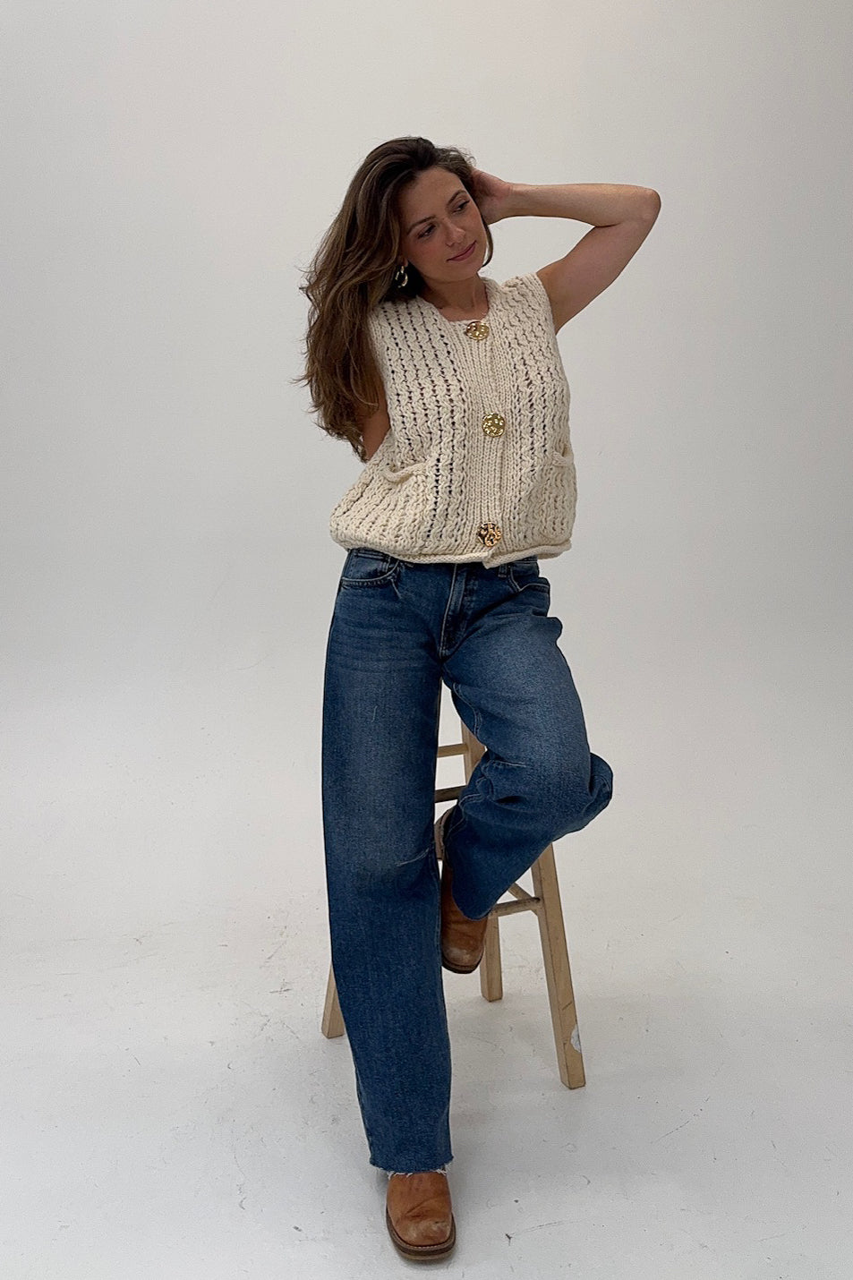 Woman wearing a beige knitted sweater and blue jeans sitting on a stool against a white background