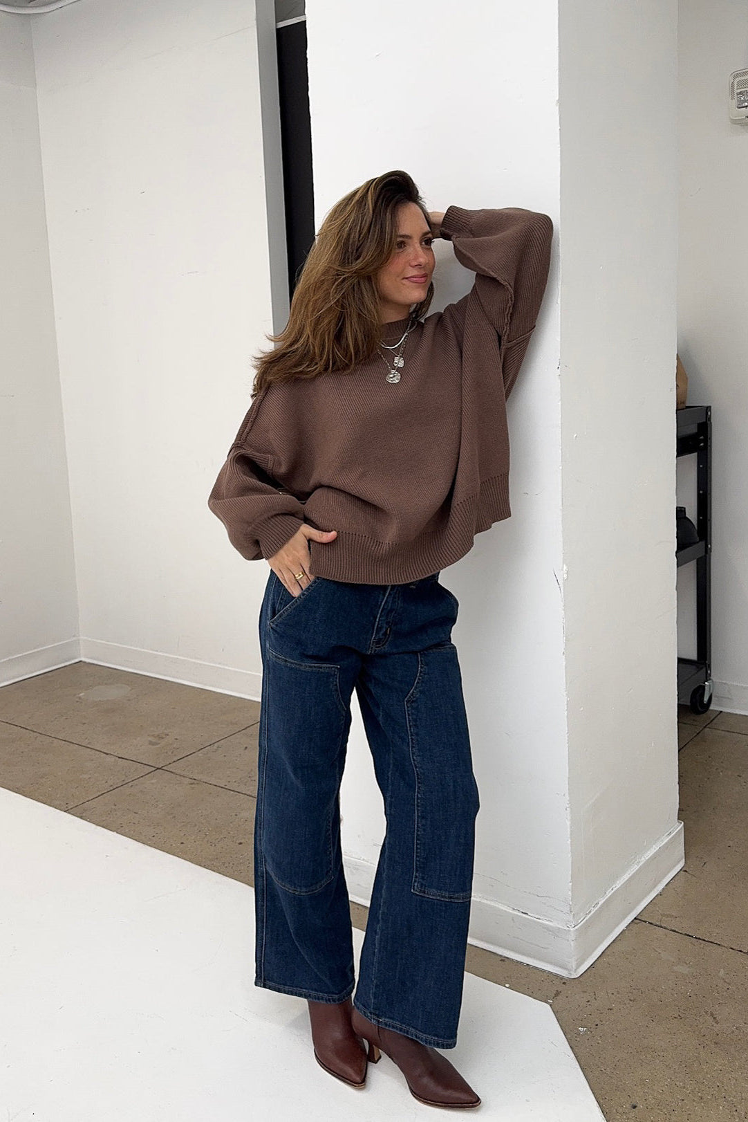 Woman wearing a brown oversized sweater and blue jeans standing against a white wall.