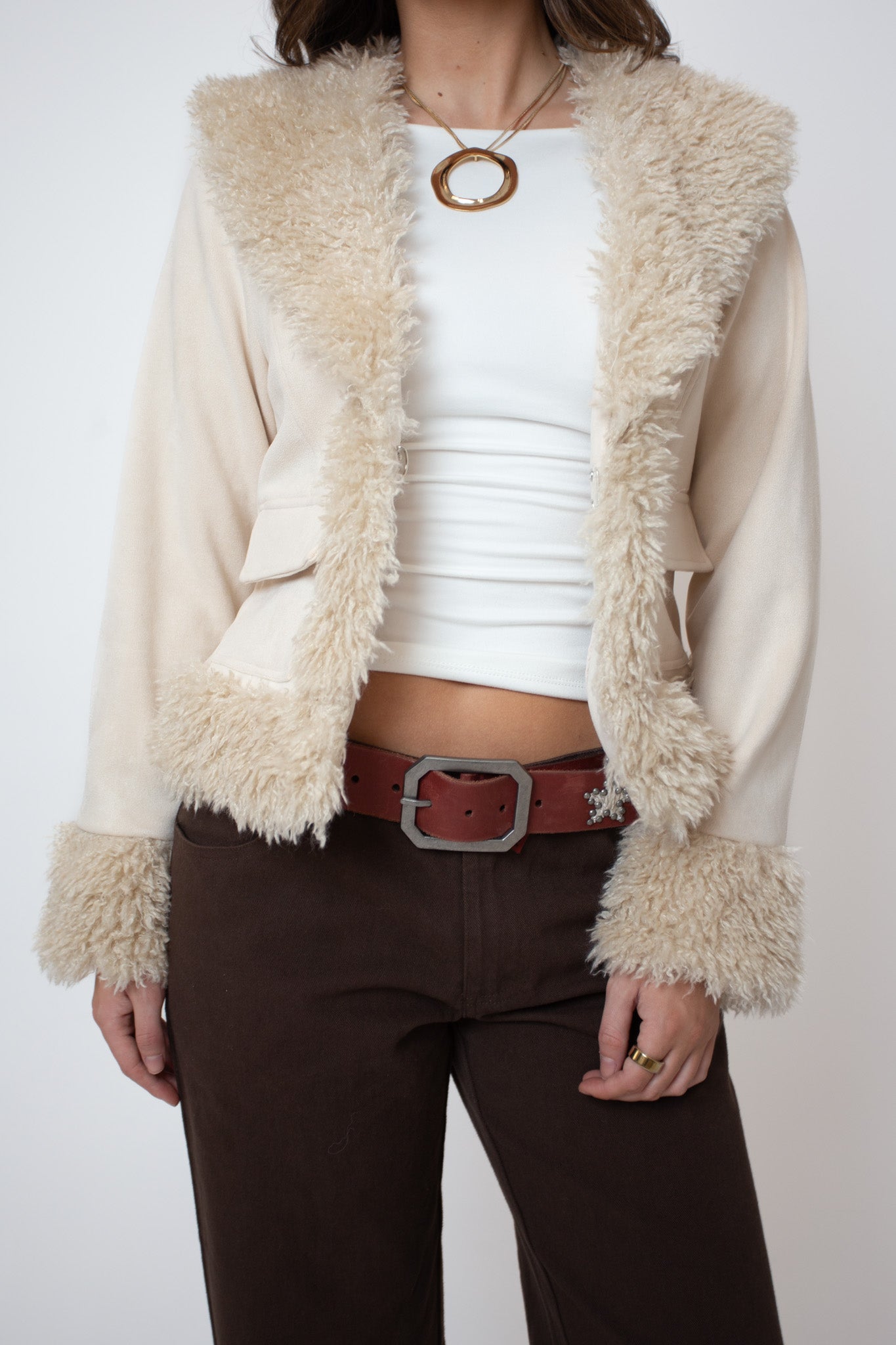Person wearing a cream-colored cardigan with fur trim, white top, brown pants, and belt on a white background