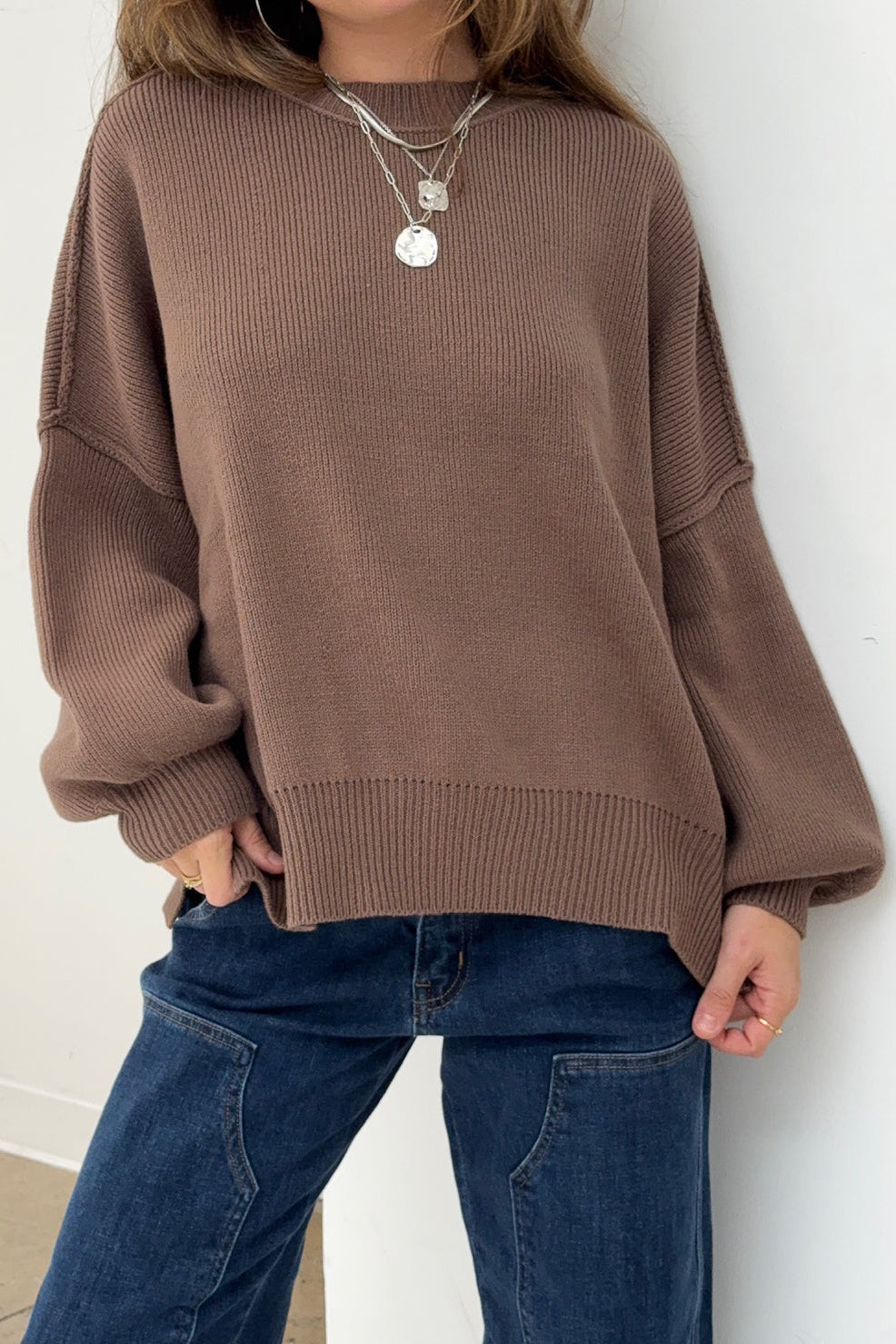 Person wearing a brown sweater and blue jeans against a white wall