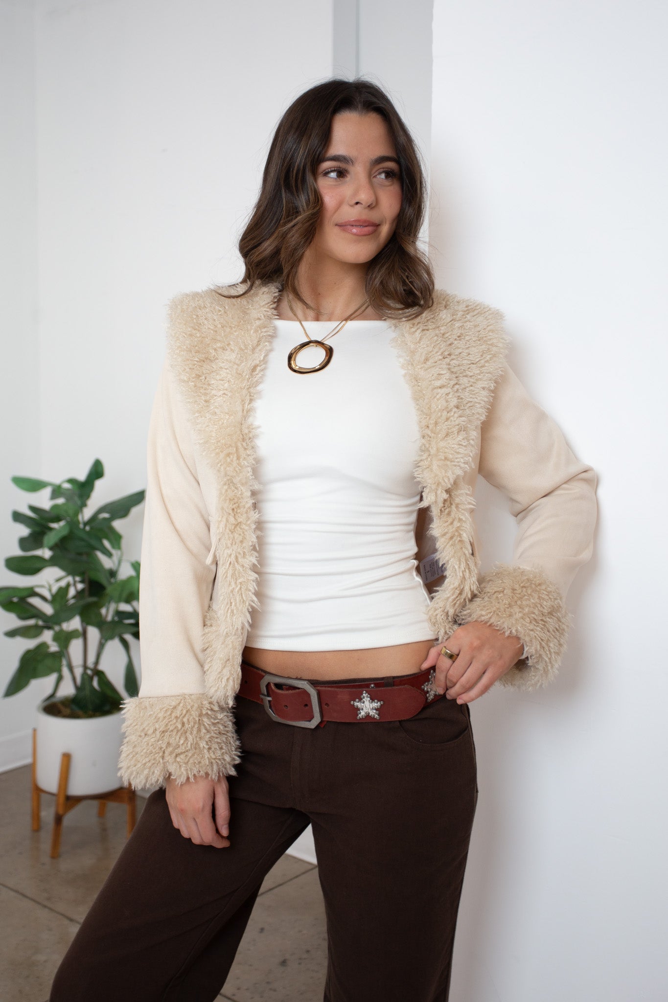 Woman wearing a beige fluffy cardigan over a white top and brown pants, standing against a white wall with a plant in the background.