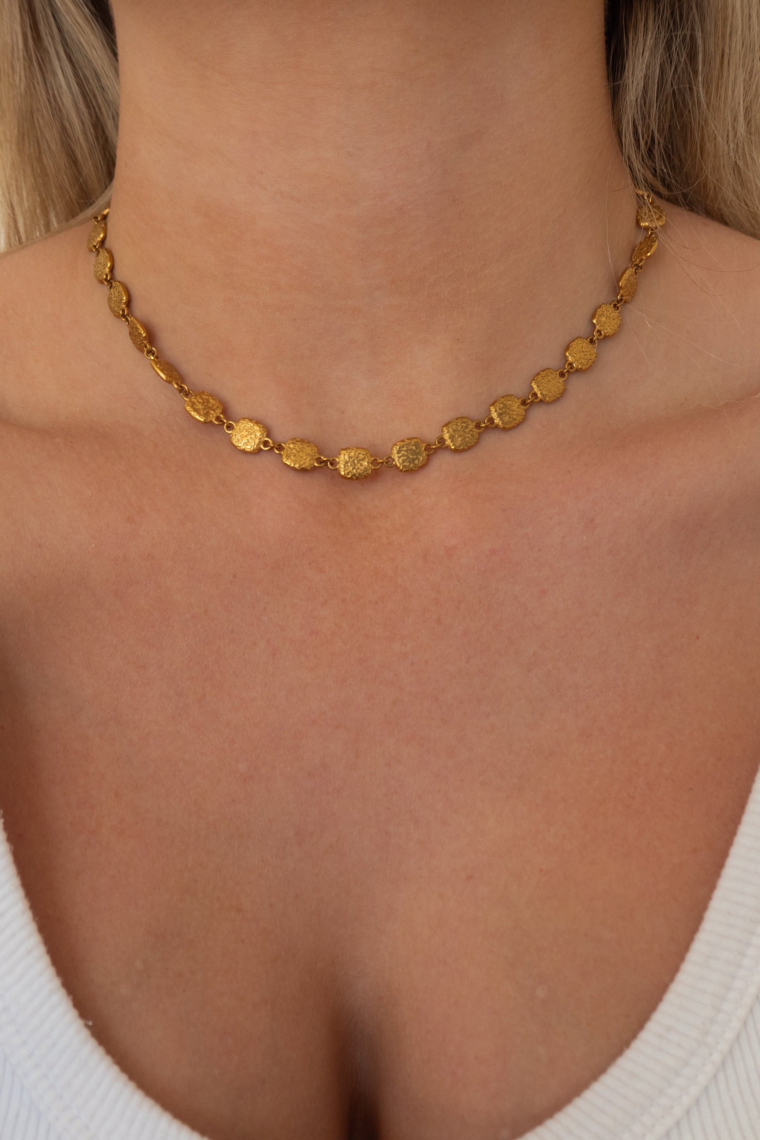 A close-up of a model's neck and upper chest wearing a delicate gold disc-link chain necklace.