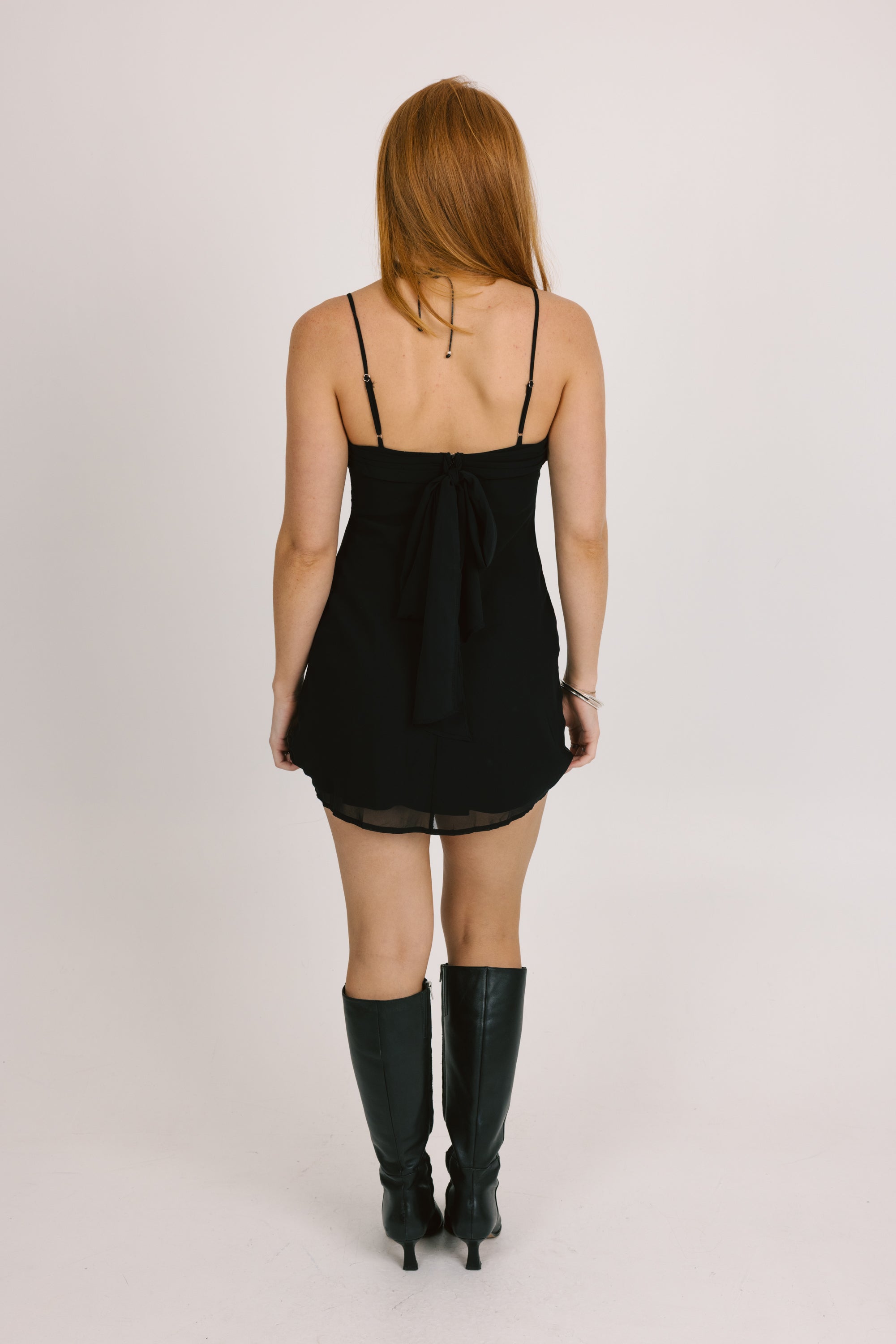 Person wearing a black dress with thin straps and black boots on a light gray background