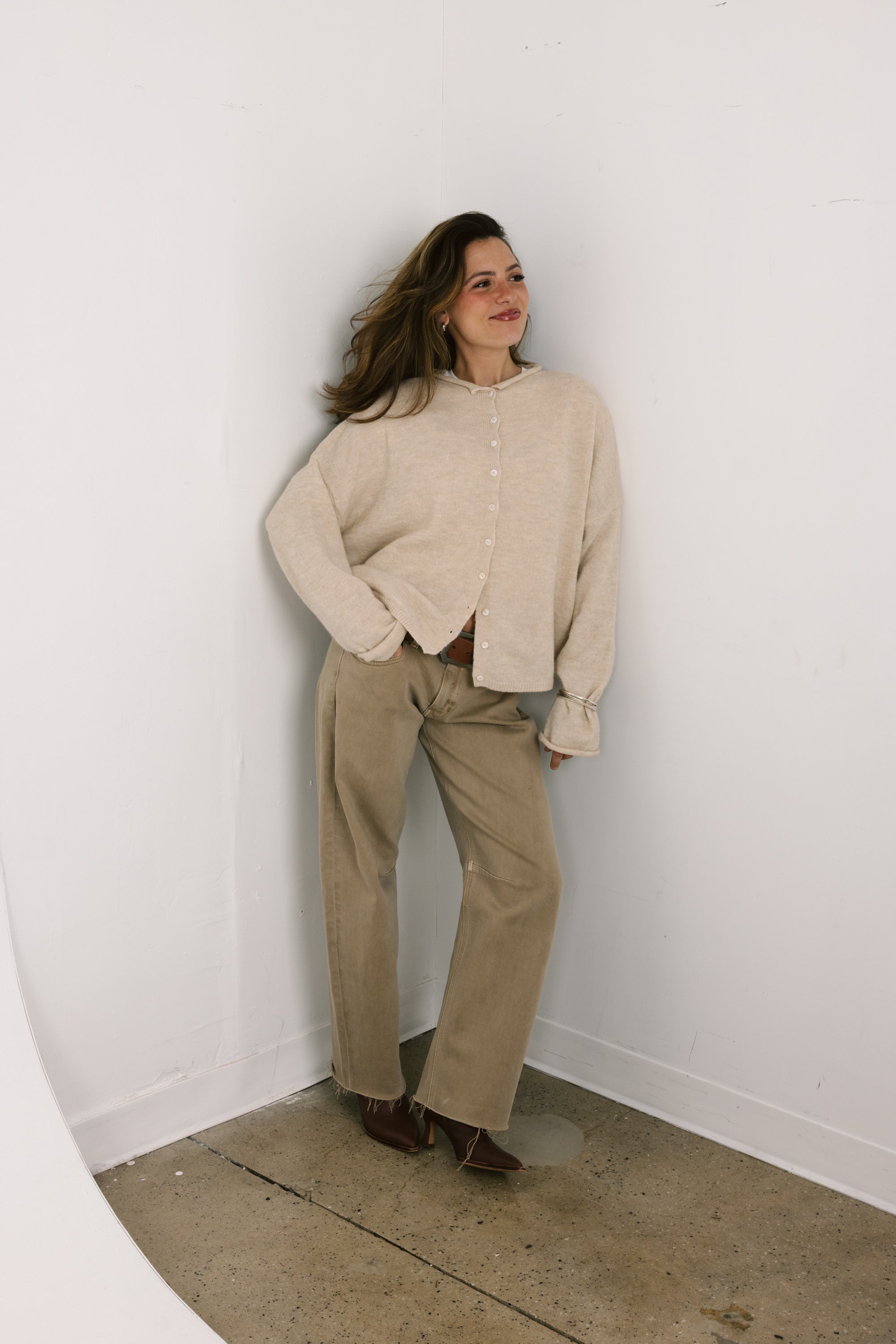 Woman wearing a beige shirt and pants standing against a white wall.