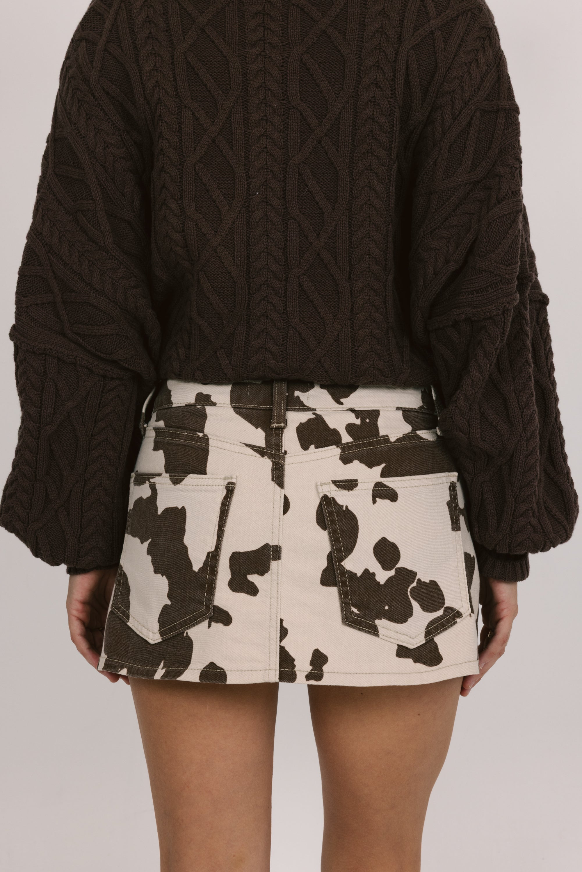 Person wearing a dark brown cable knit sweater and a patterned skirt on a plain background