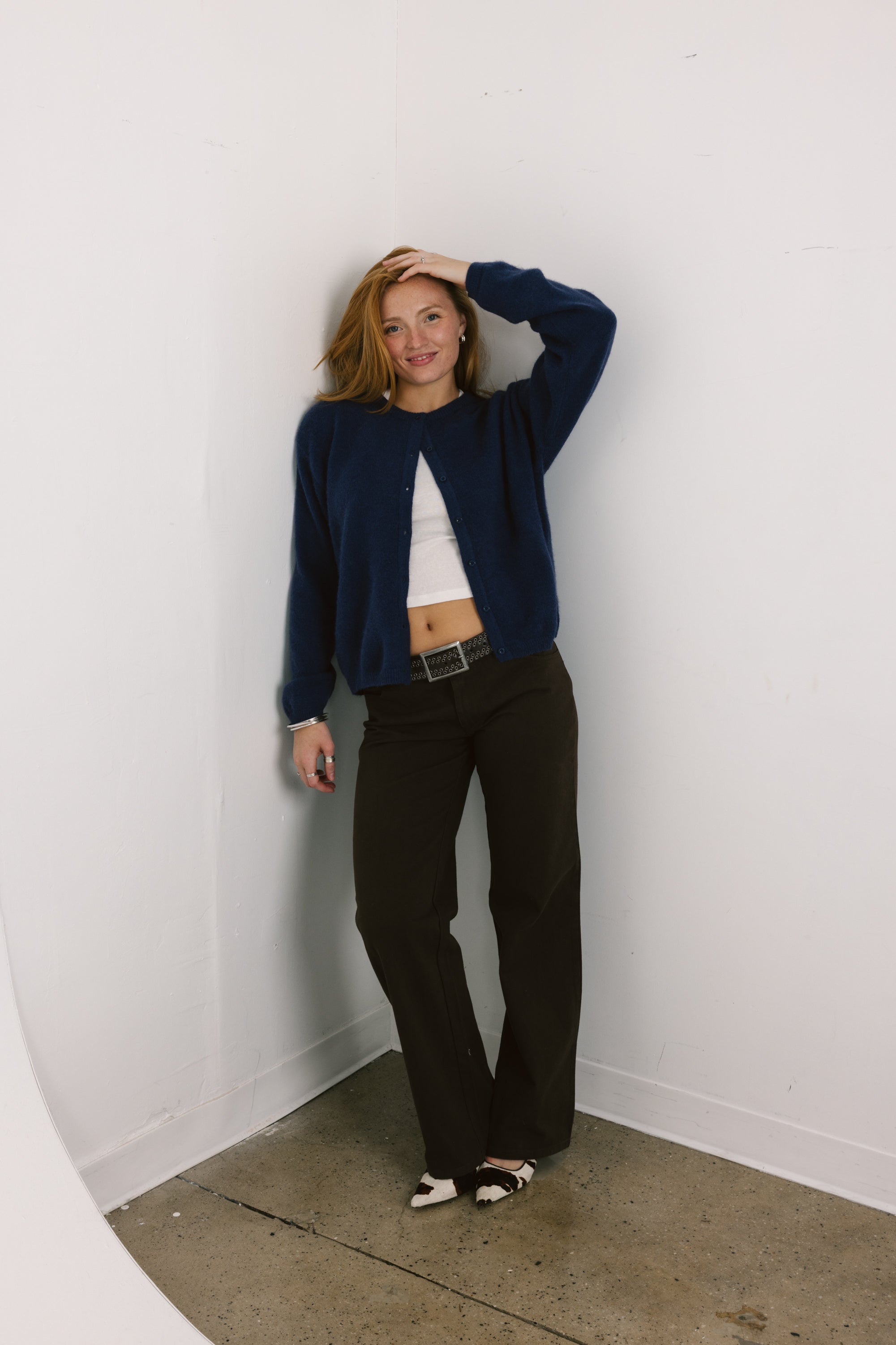 Woman wearing a navy cardigan, white top, and black pants against a white wall.
