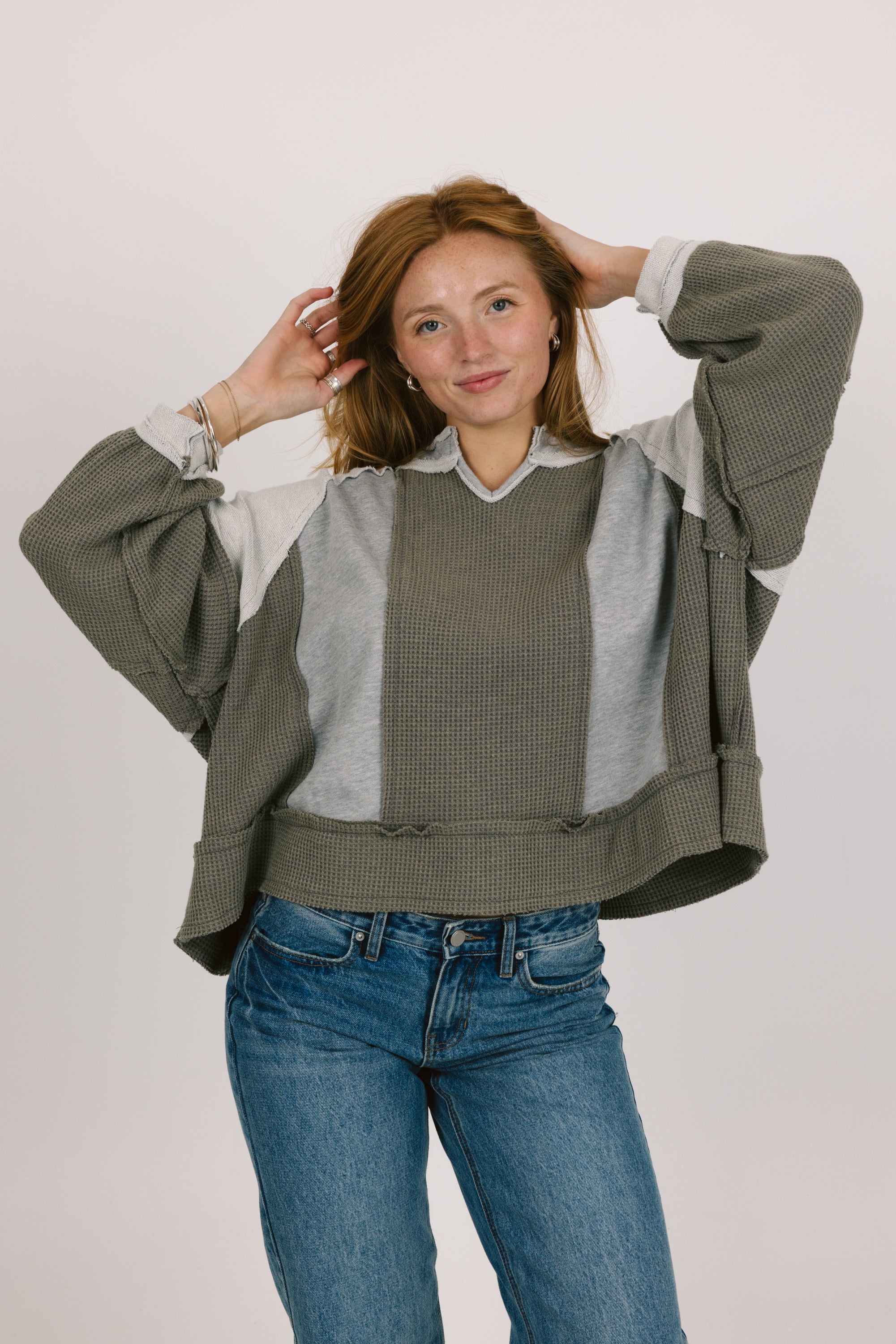 Woman wearing a layered outfit with a green jacket, gray shirt, and blue jeans on a plain background
