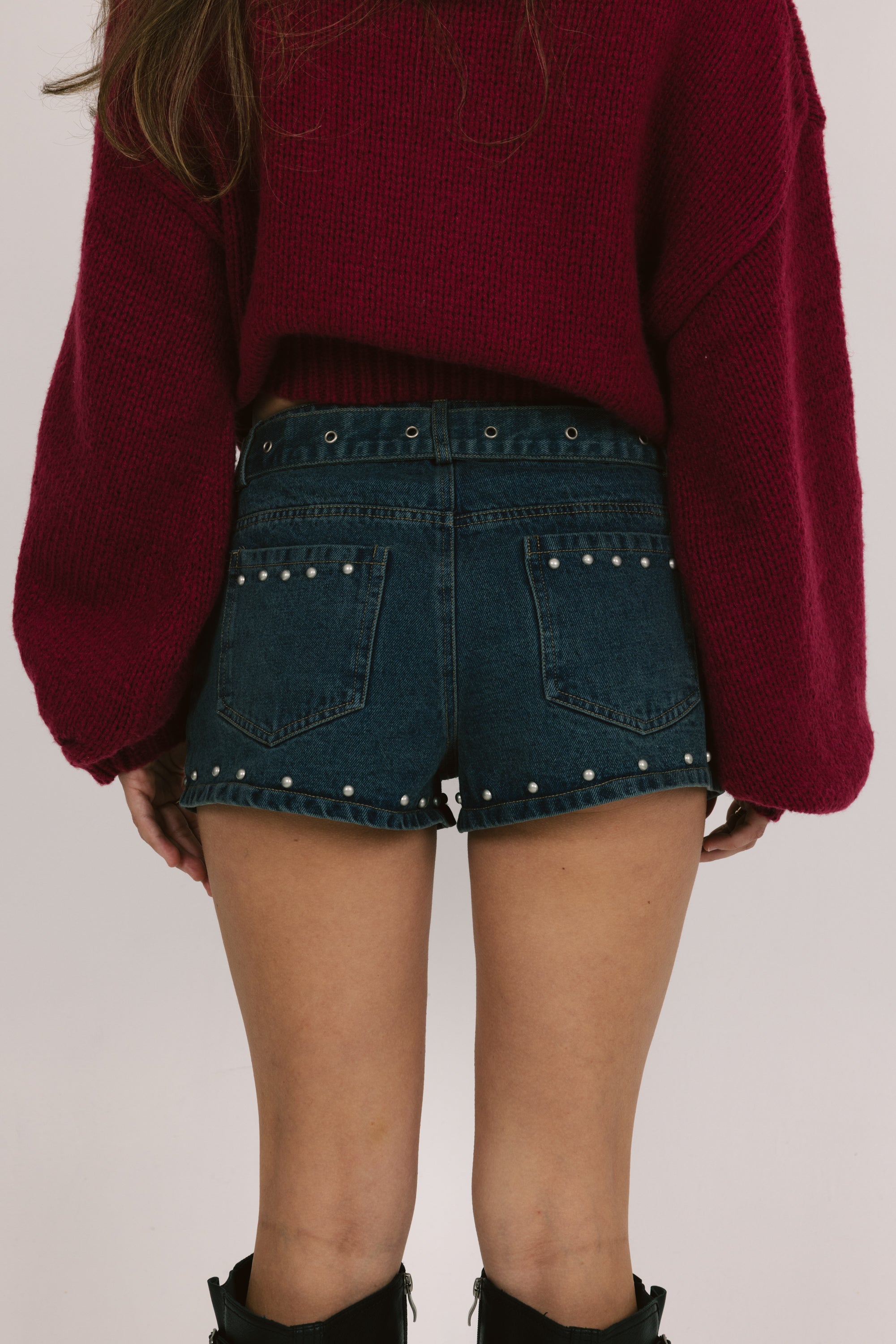 Person wearing a burgundy sweater and denim shorts with pearl details on a plain background