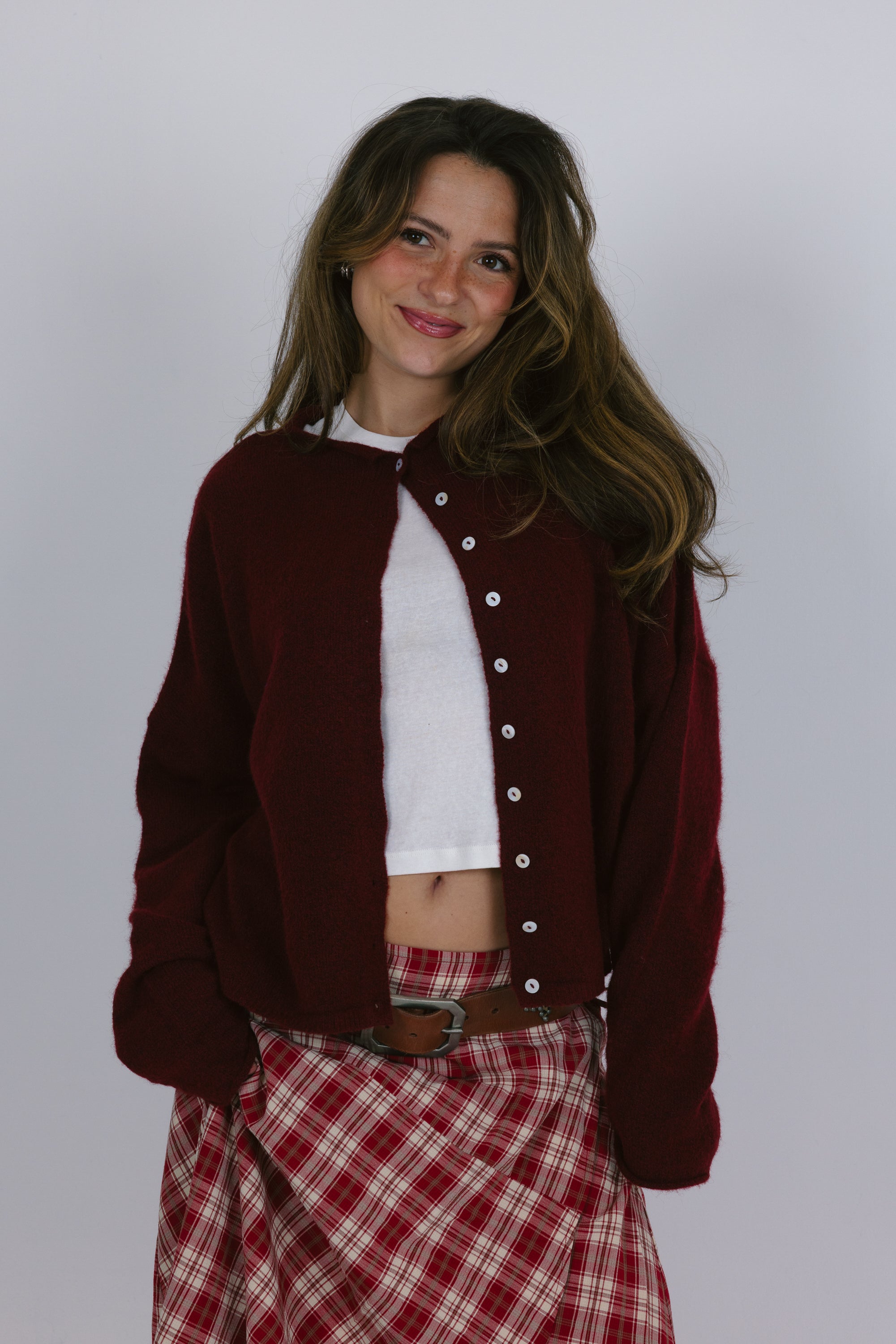 Woman wearing a burgundy cardigan over a white shirt and red plaid skirt on a plain background