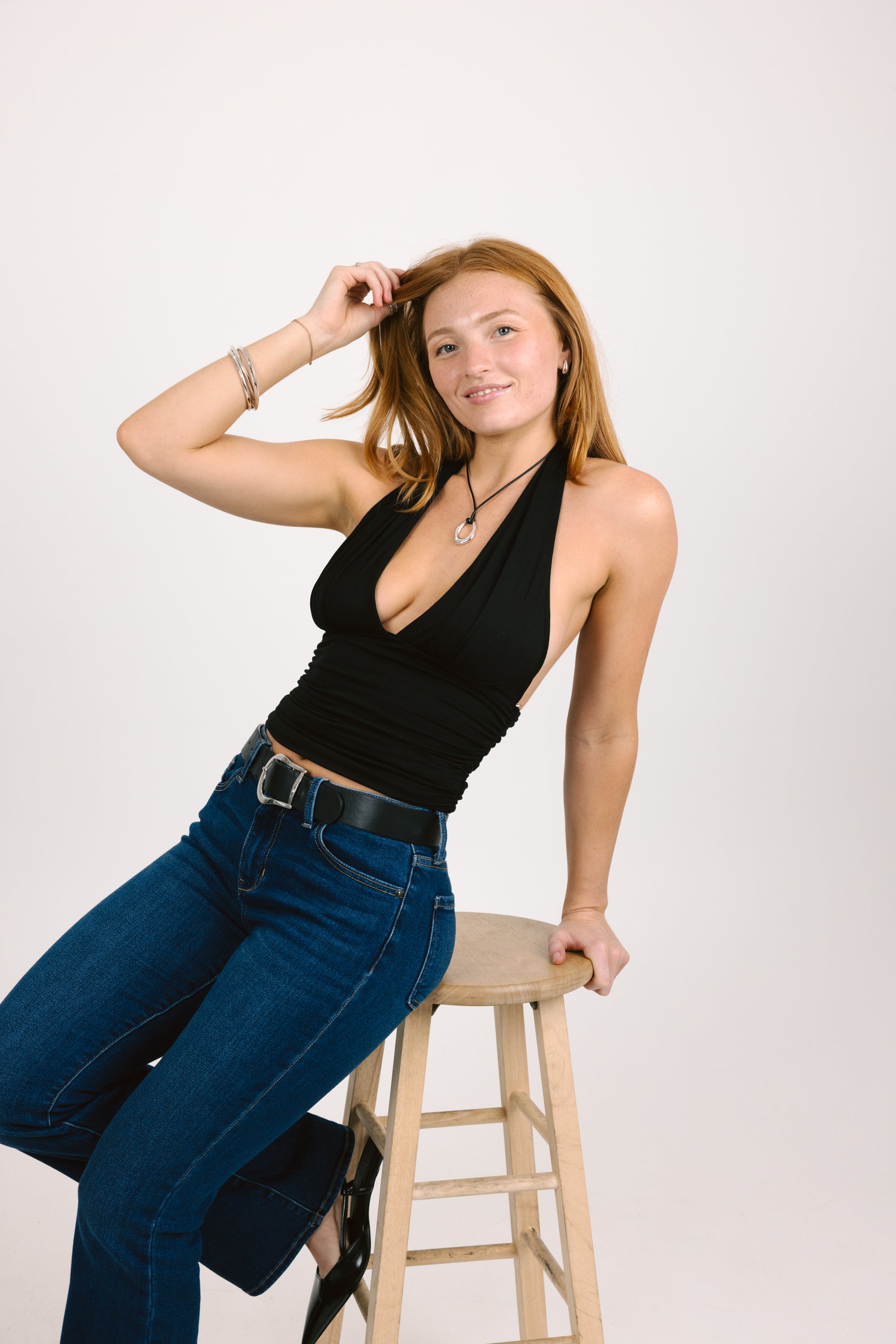 Woman wearing a black top and blue jeans sitting on a wooden stool against a white background