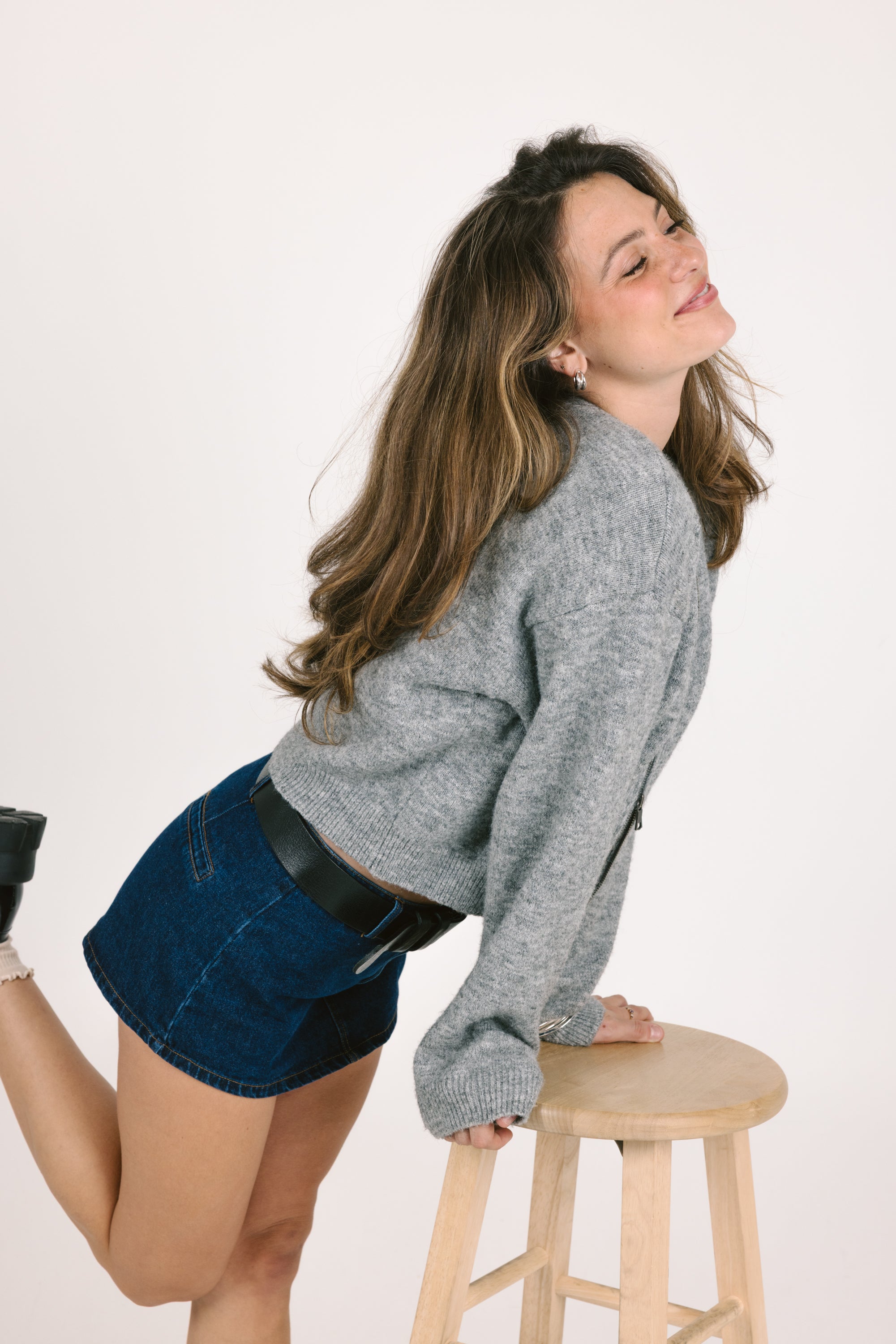 Woman in a gray sweater and denim shorts posing on a stool against a white background