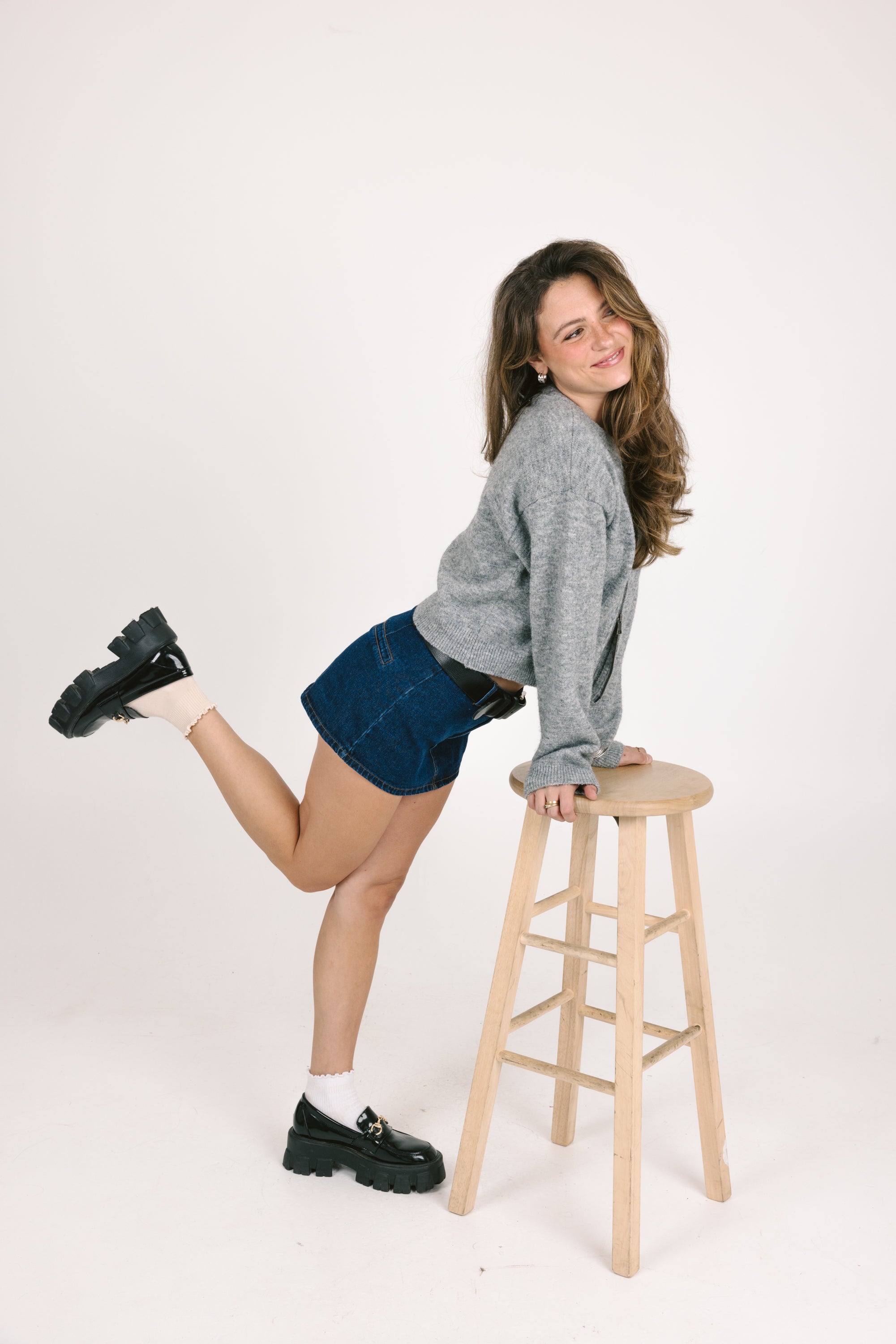 Woman sitting on a wooden stool wearing a gray sweater and denim shorts against a white background