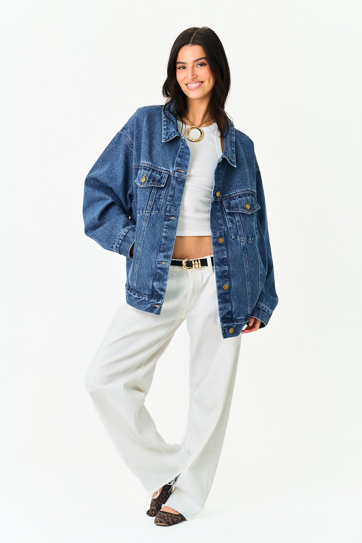 A woman modeling an oversized blue denim jacket over a white crop top and white wide-leg pants, standing against a plain white studio background.