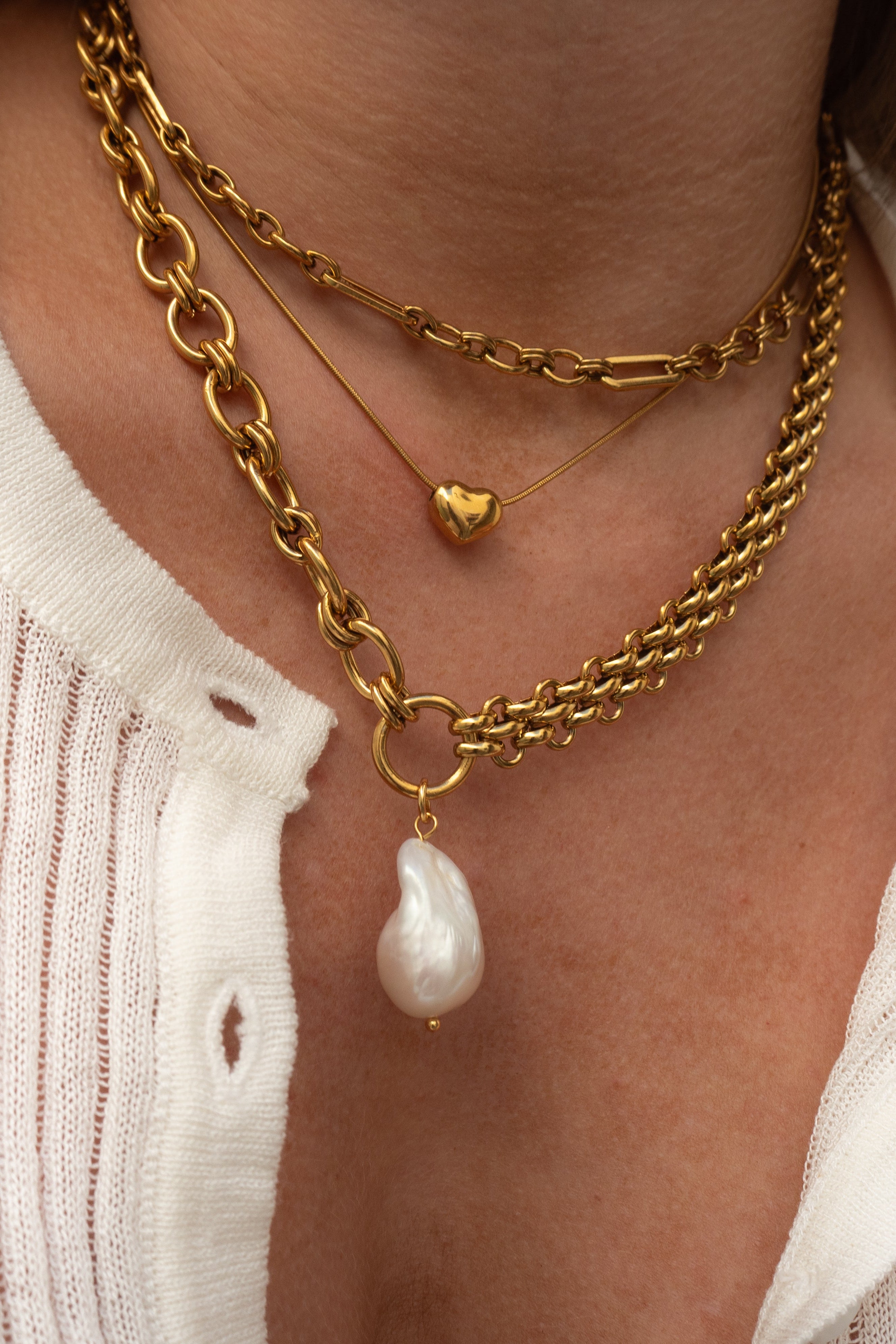 Close-up of a person wearing gold chain necklaces with a heart pendant and a pearl pendant.