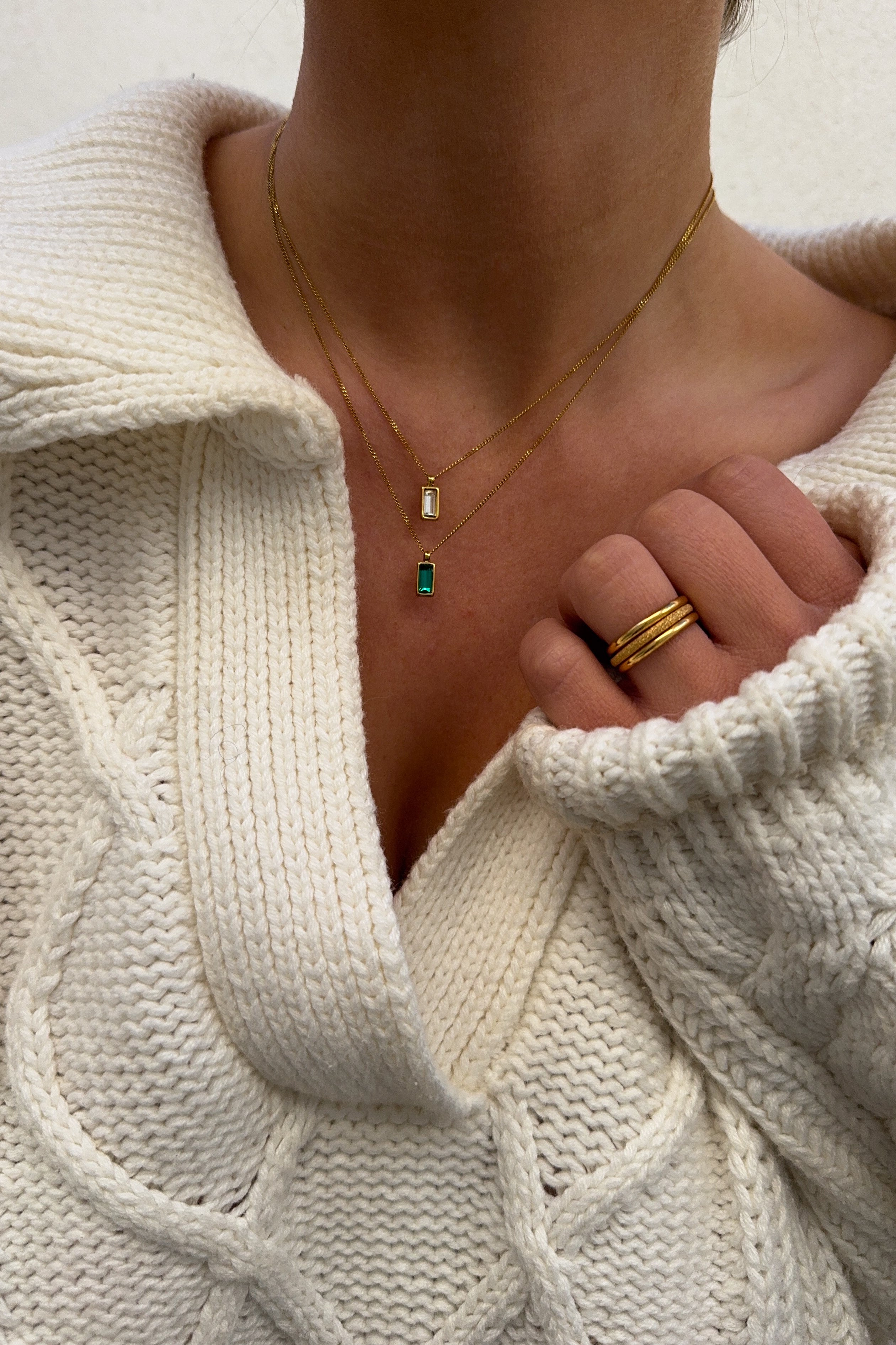 Close-up of a person wearing a gold necklace with an emerald pendant and gold rings, with a white sweater partially visible.