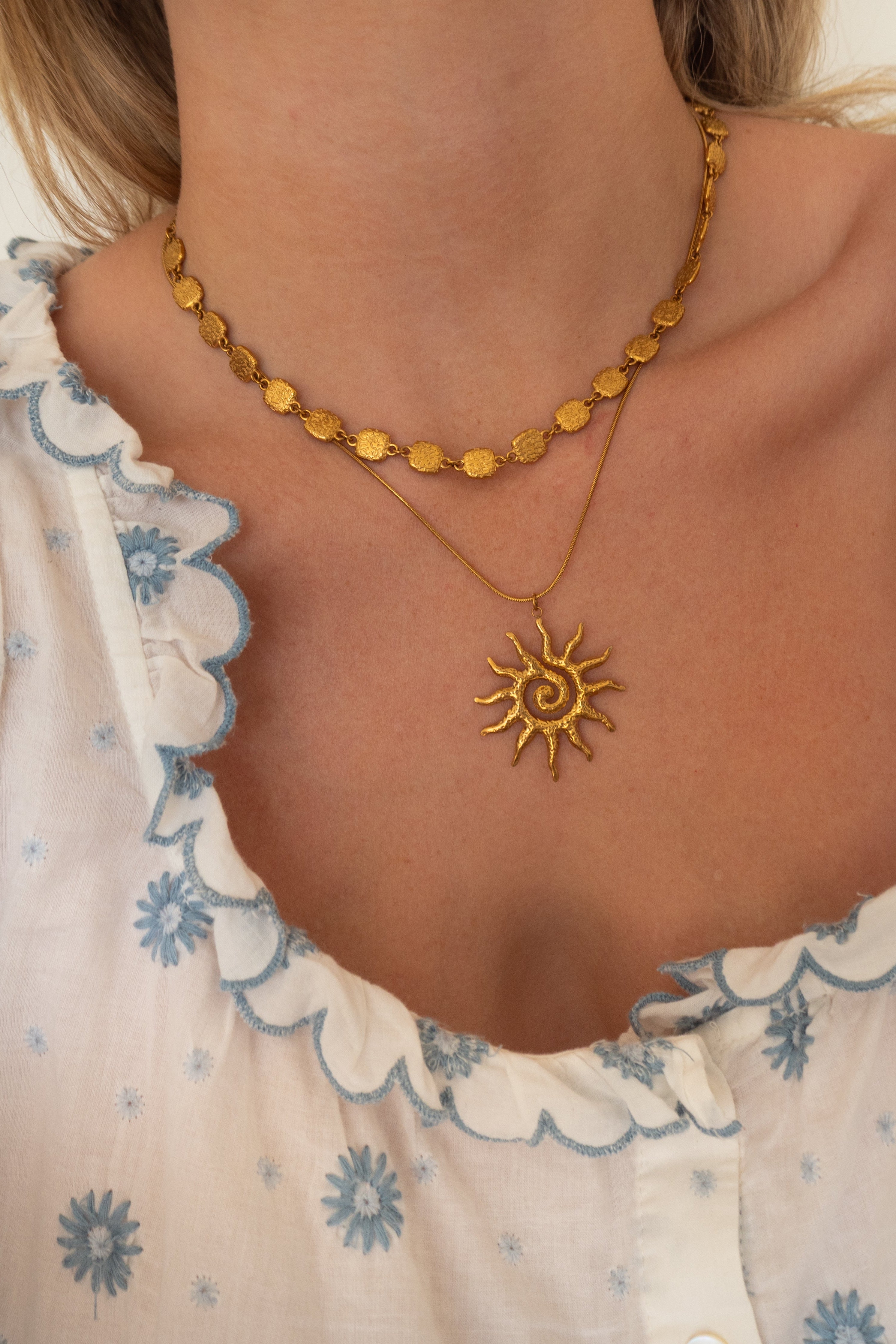A close-up of a person's neck and chest wearing two gold necklaces: a shorter chain with flat circular links and a longer chain with a sun pendant, styled with a white ruffled blouse featuring blue floral patterns.