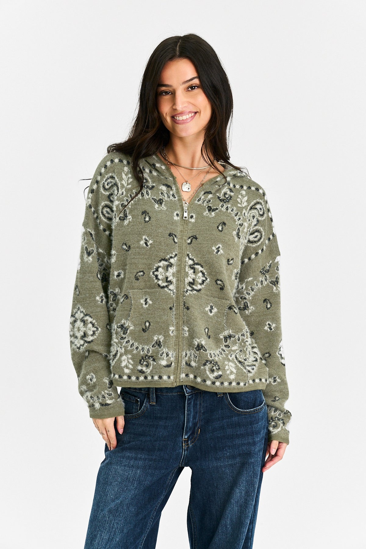 A woman modeling a sage green zip-up hoodie with a black and white paisley bandana pattern, paired with dark blue jeans, against a plain light gray background.