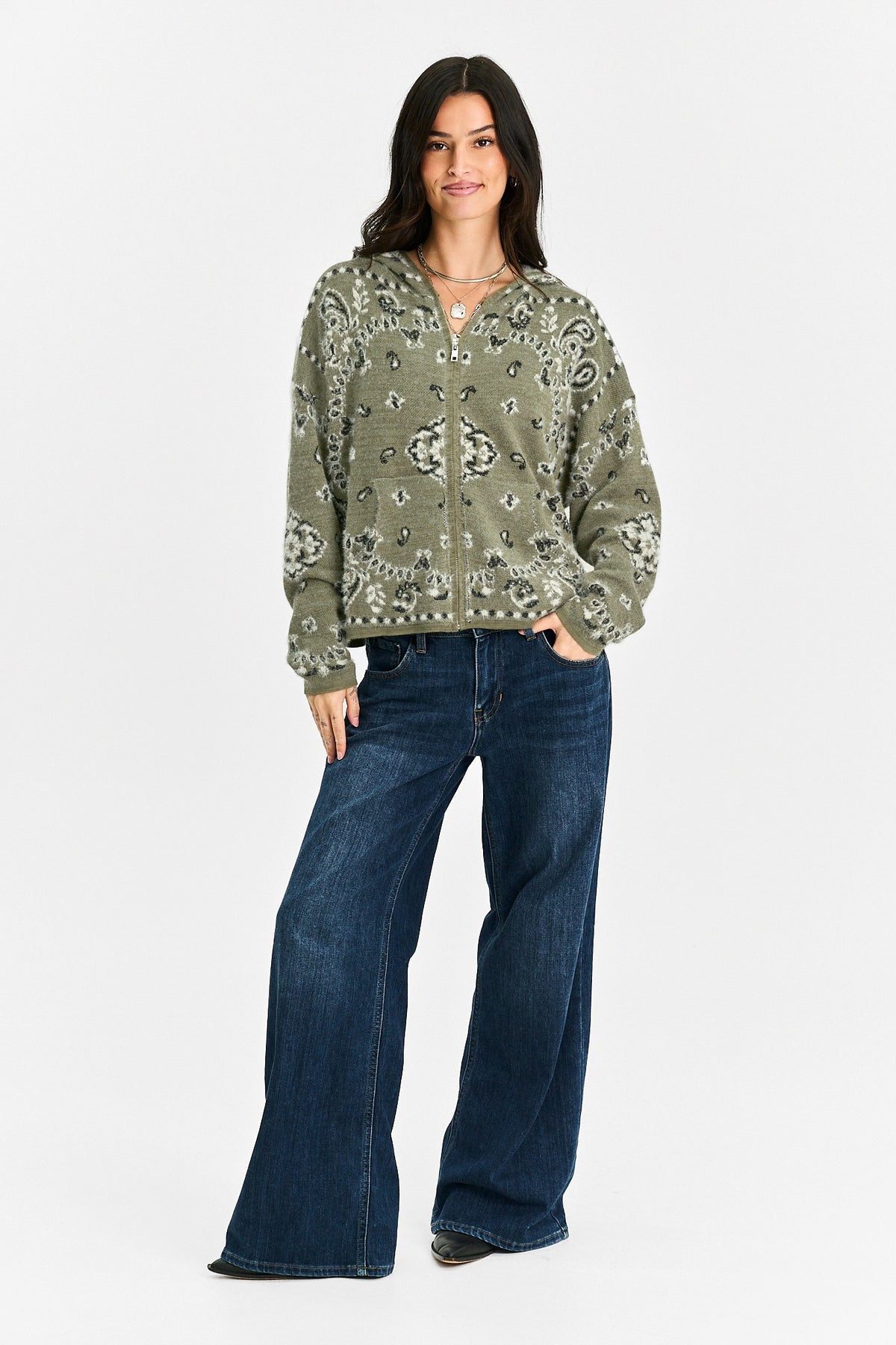 A woman modeling a sage green zip-up hoodie with a white paisley pattern, paired with dark blue wide-leg jeans, against a plain light gray background.