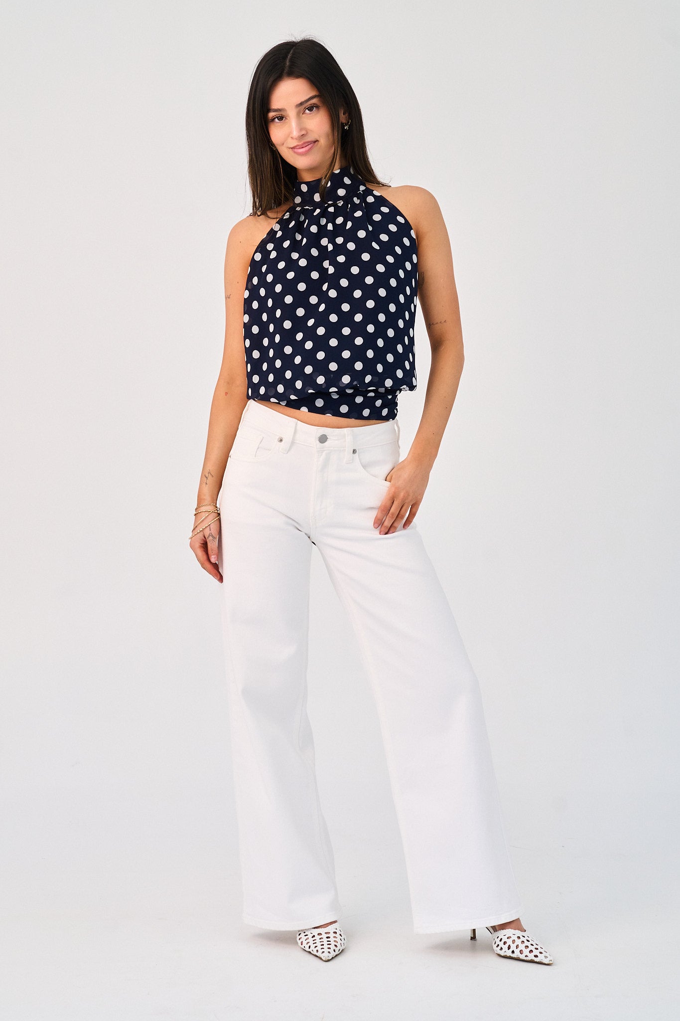 Woman wearing a black polka dot halter top and white pants on a white background