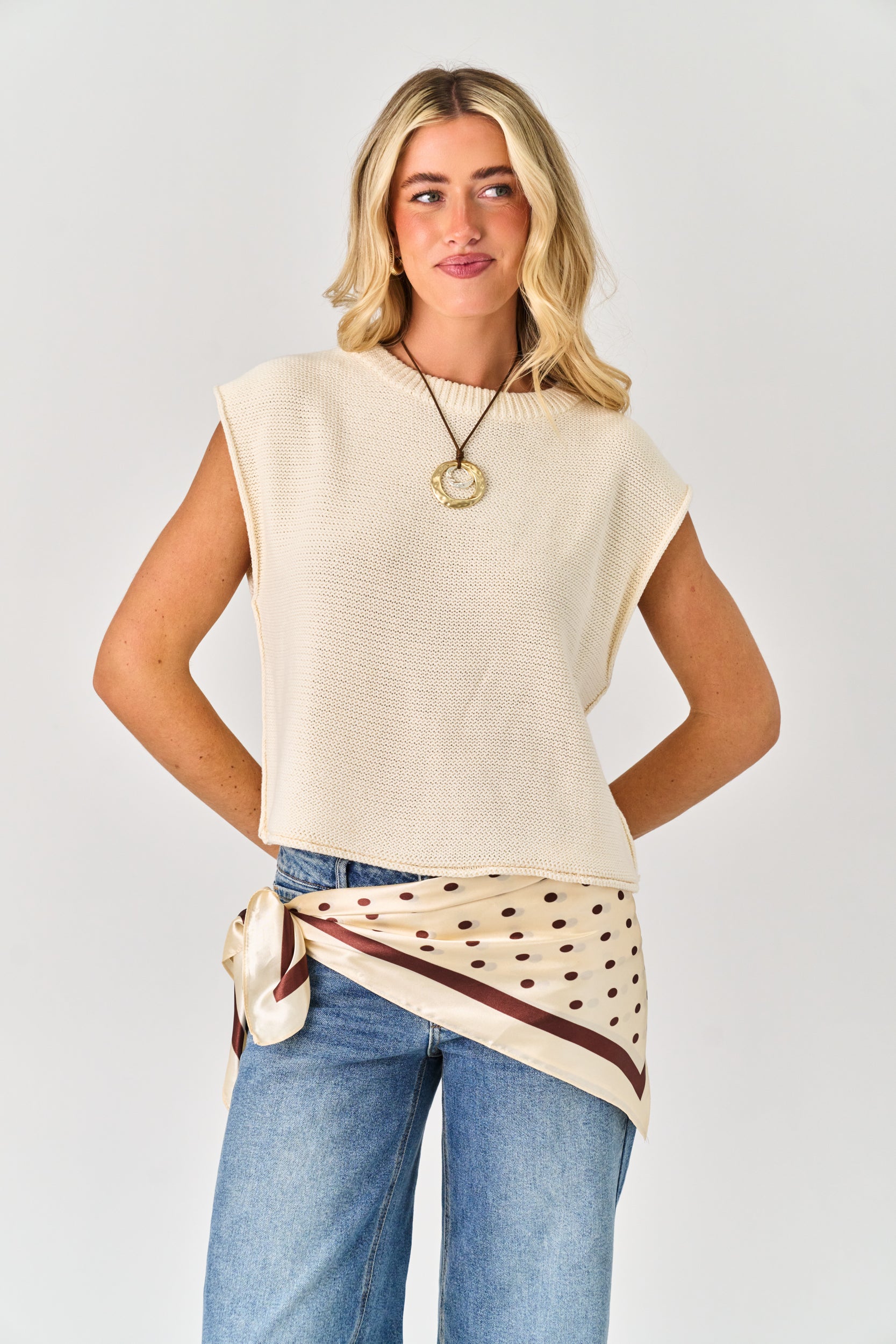 Woman wearing a beige sleeveless top with a polka dot scarf, standing against a plain background