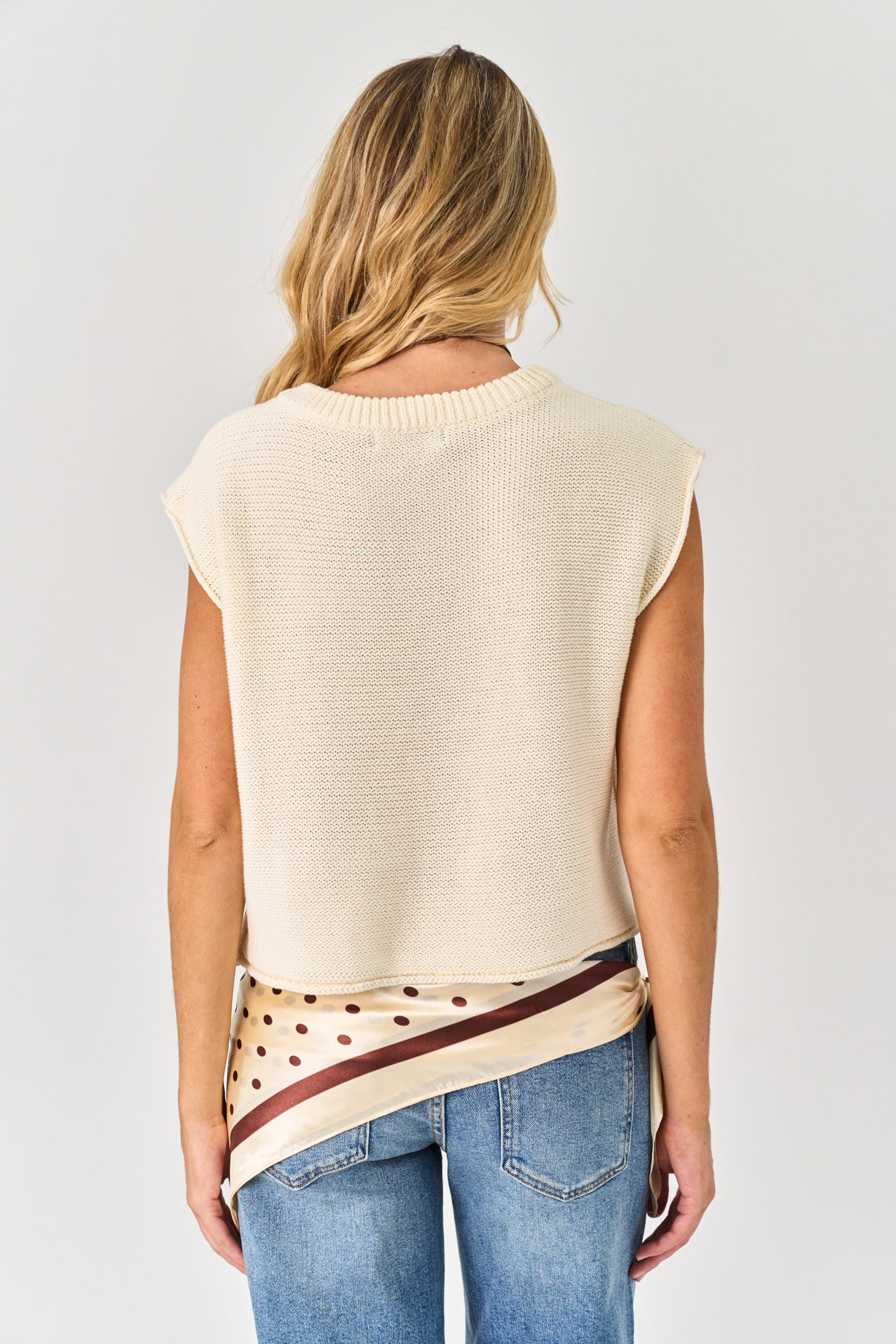 Person wearing a beige sleeveless top with a patterned belt and blue jeans on a plain background