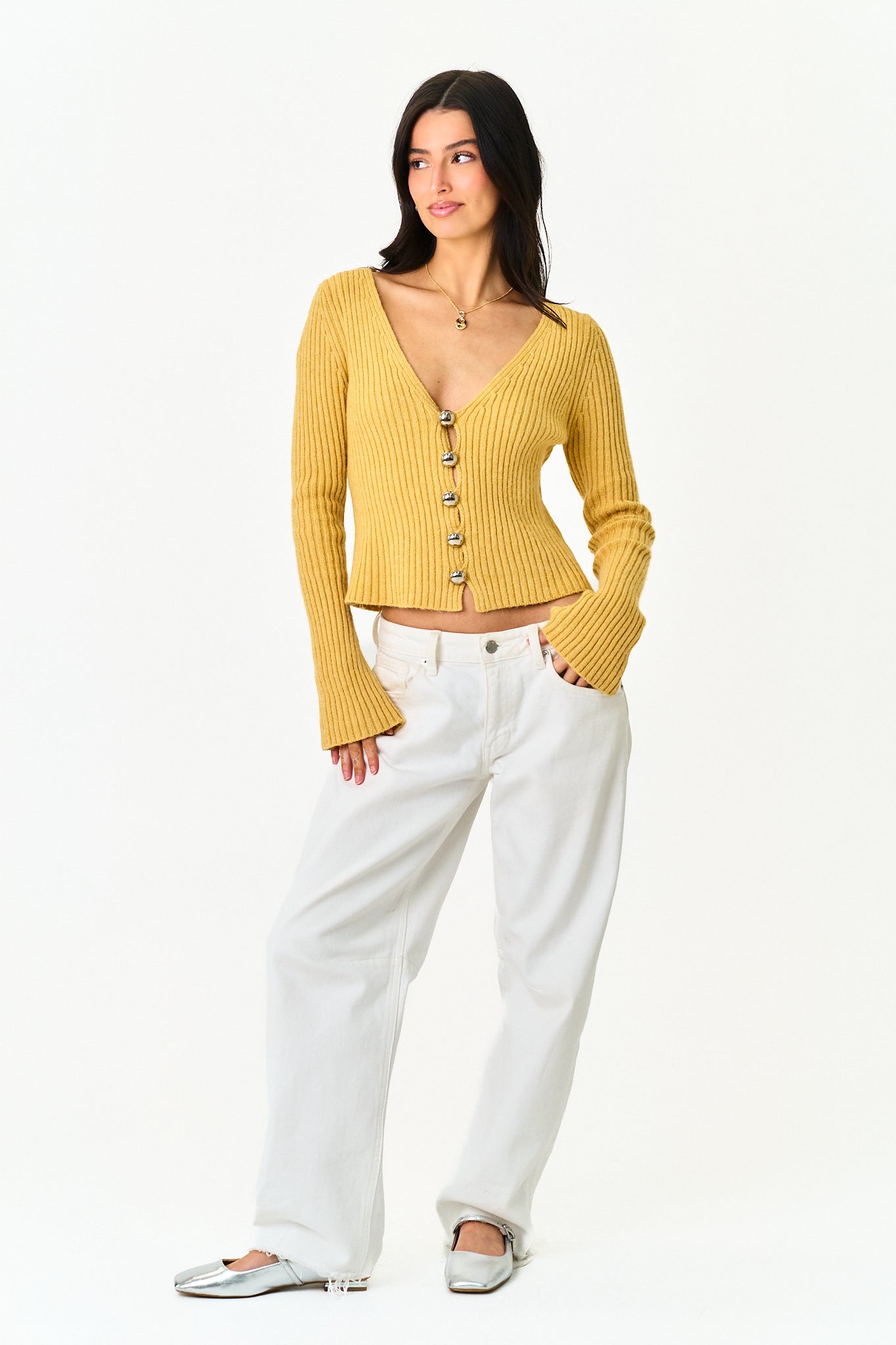 A woman modeling a mustard yellow ribbed cardigan with a button front and flared sleeves, paired with white wide-leg pants and silver flats, against a plain white background.