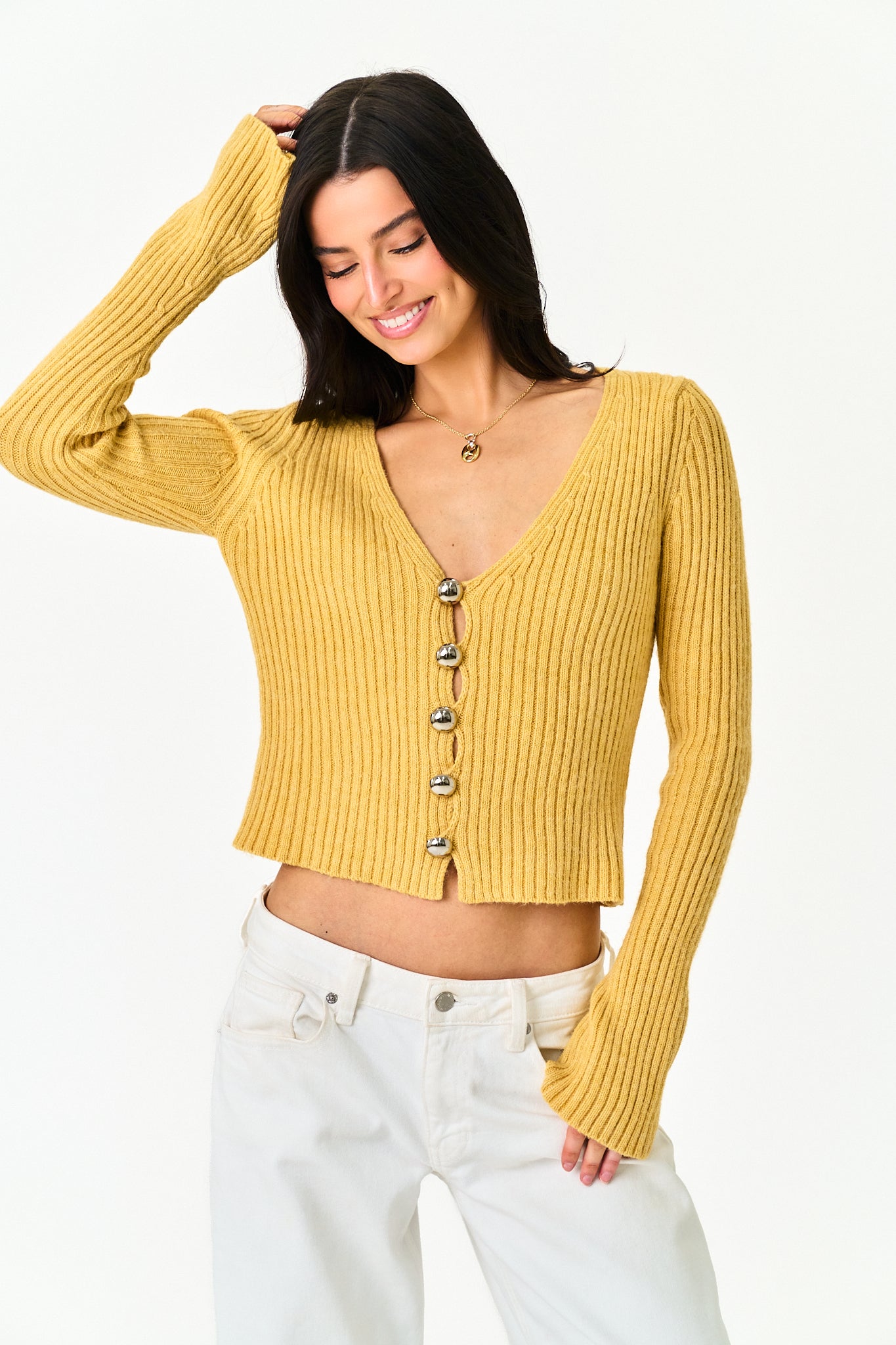 A woman wearing a ribbed mustard yellow long-sleeve cardigan with a V-neck and front buttons, paired with white pants, against a plain white background.