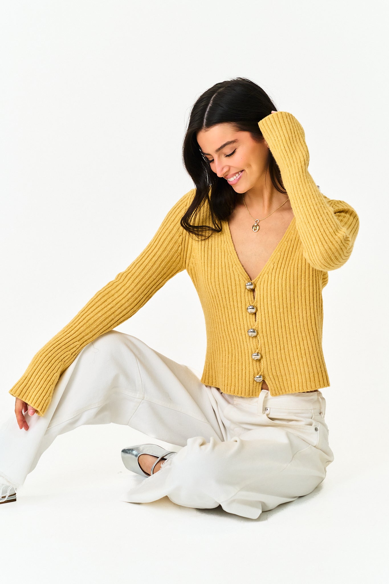 A woman sitting on a white studio floor wearing a mustard yellow ribbed knit cardigan with silver buttons, white wide-leg pants, and silver pointed shoes.