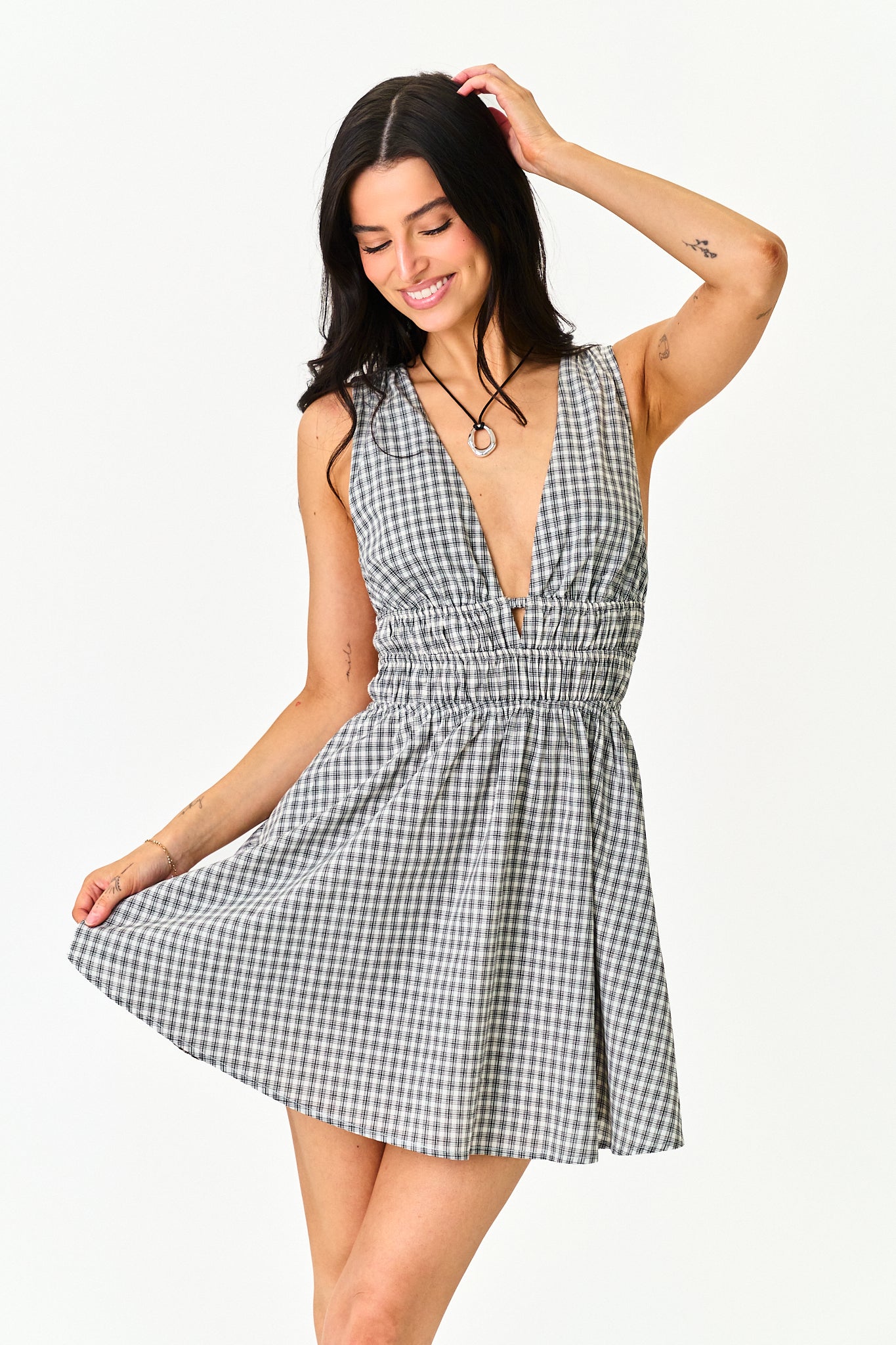 A woman modeling a sleeveless black and white gingham mini dress with a smocked waist and a deep V-neckline, set against a plain white background.