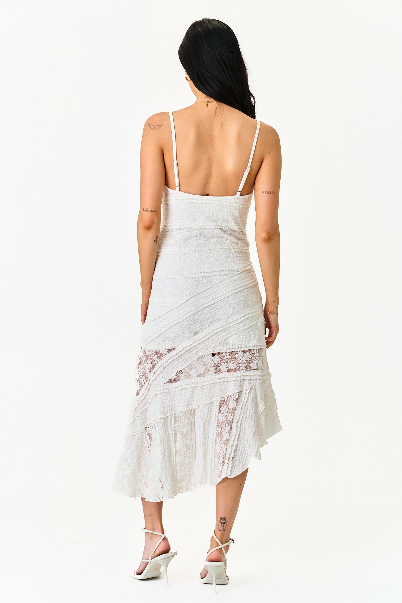 A woman shown from the back wearing a white spaghetti-strap dress with lace panels and a ruffled hem, paired with white strappy heels, against a plain white background.