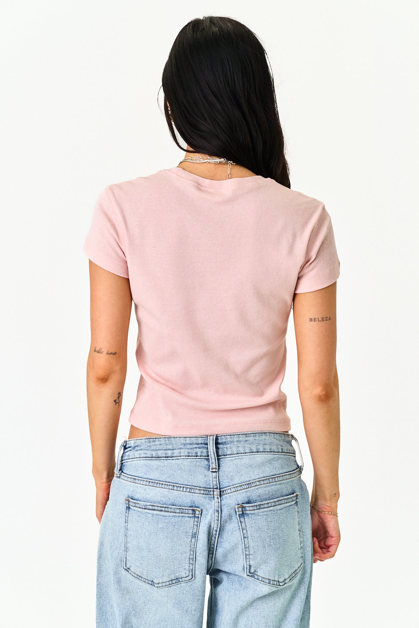 A woman viewed from behind wearing a light pink short-sleeve t-shirt and light blue jeans against a plain white background.