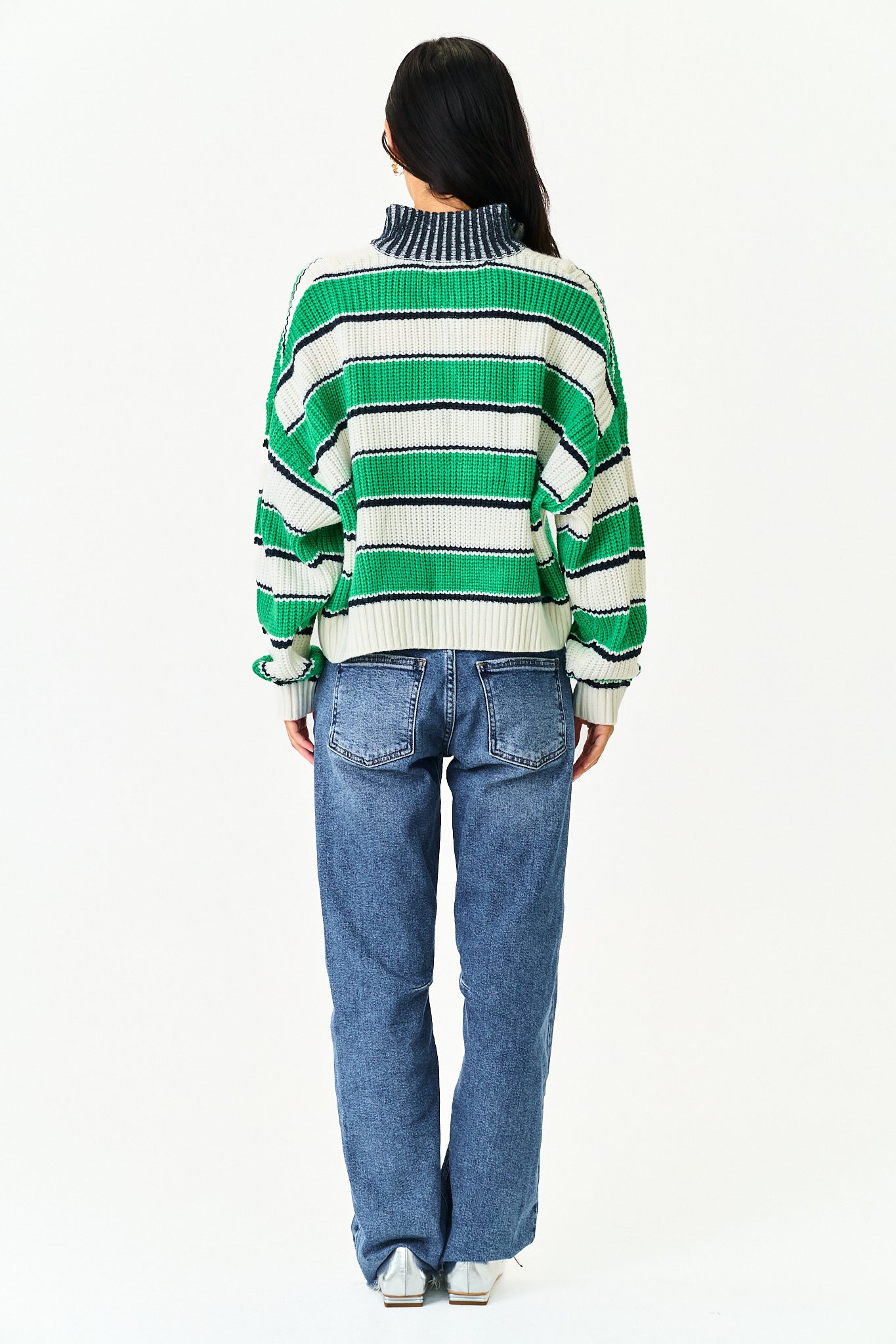 A model viewed from the back wearing a green and white striped knit sweater with a dark blue ribbed mock neck, paired with blue jeans, against a plain white background.