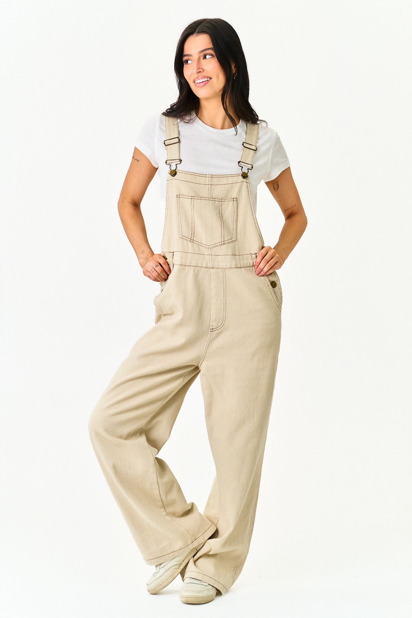A woman modeling a pair of light beige wide-leg denim bib overalls over a white t-shirt, standing against a solid white studio background.
